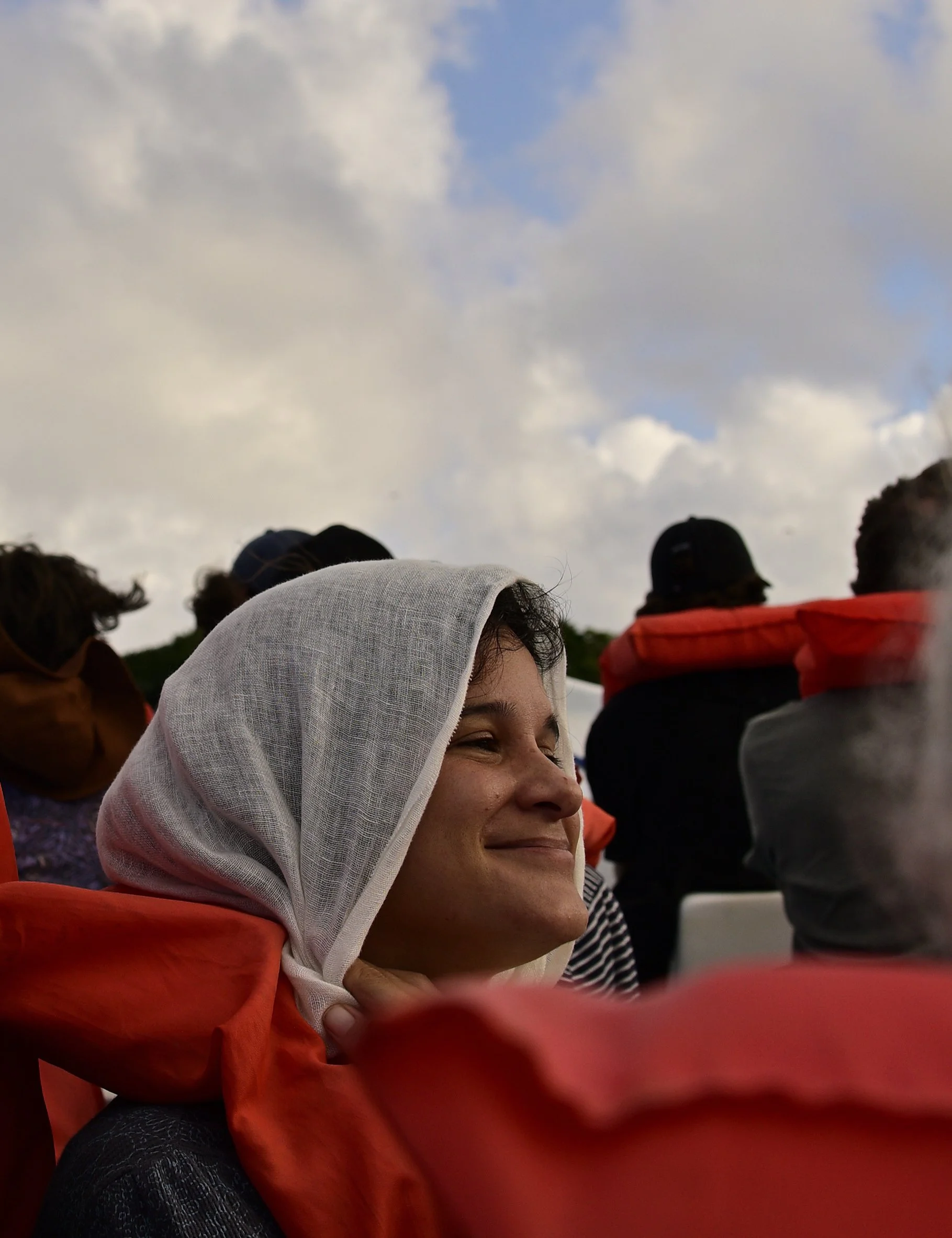 People gathered outdoors under cloudy sky, some wearing hats and headscarves, smiling and facing the same direction.