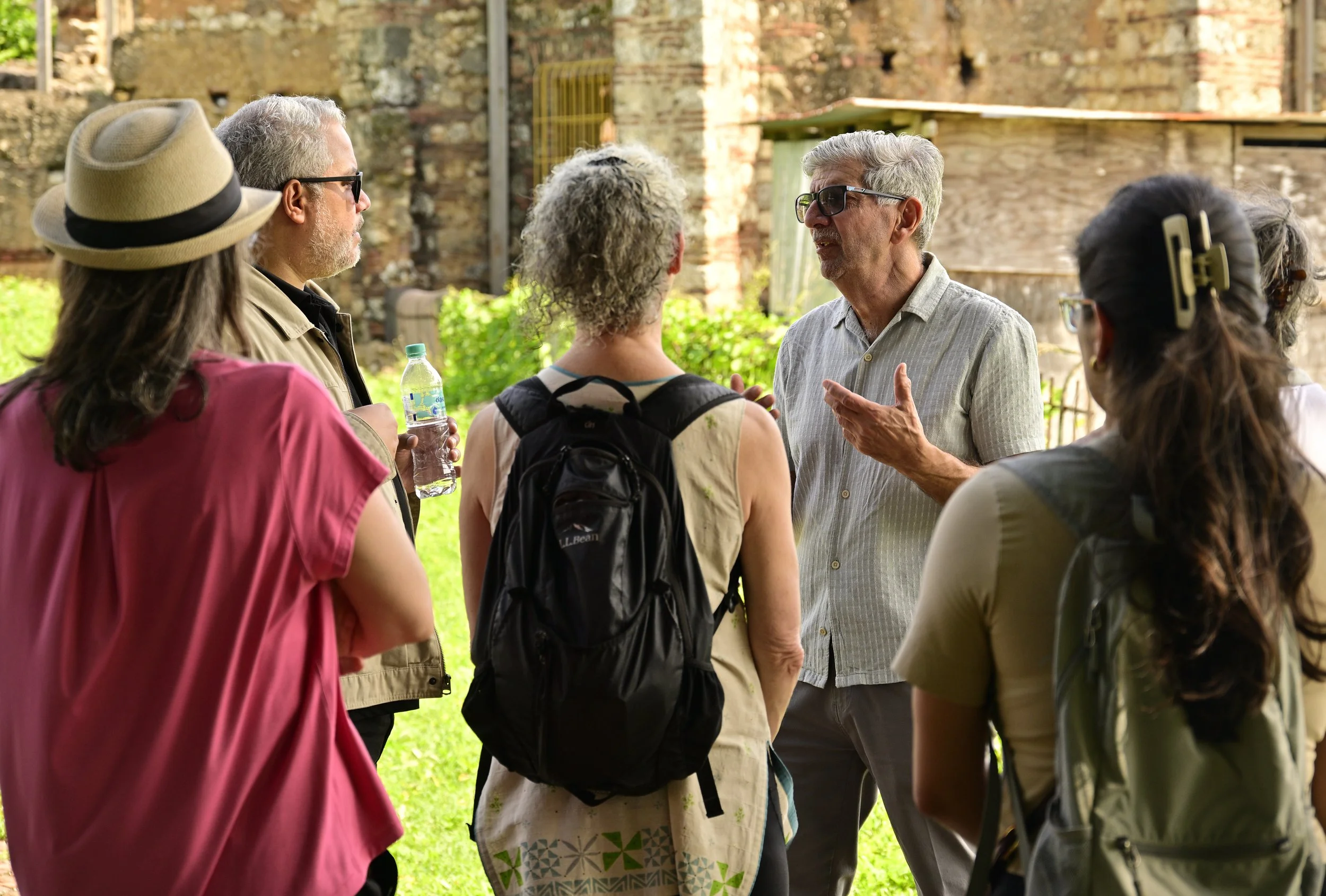 A group of people having a conversation outdoors near a weathered brick wall. The group includes men and women wearing casual clothing, hats, and sunglasses, with some carrying backpacks and a water bottle.