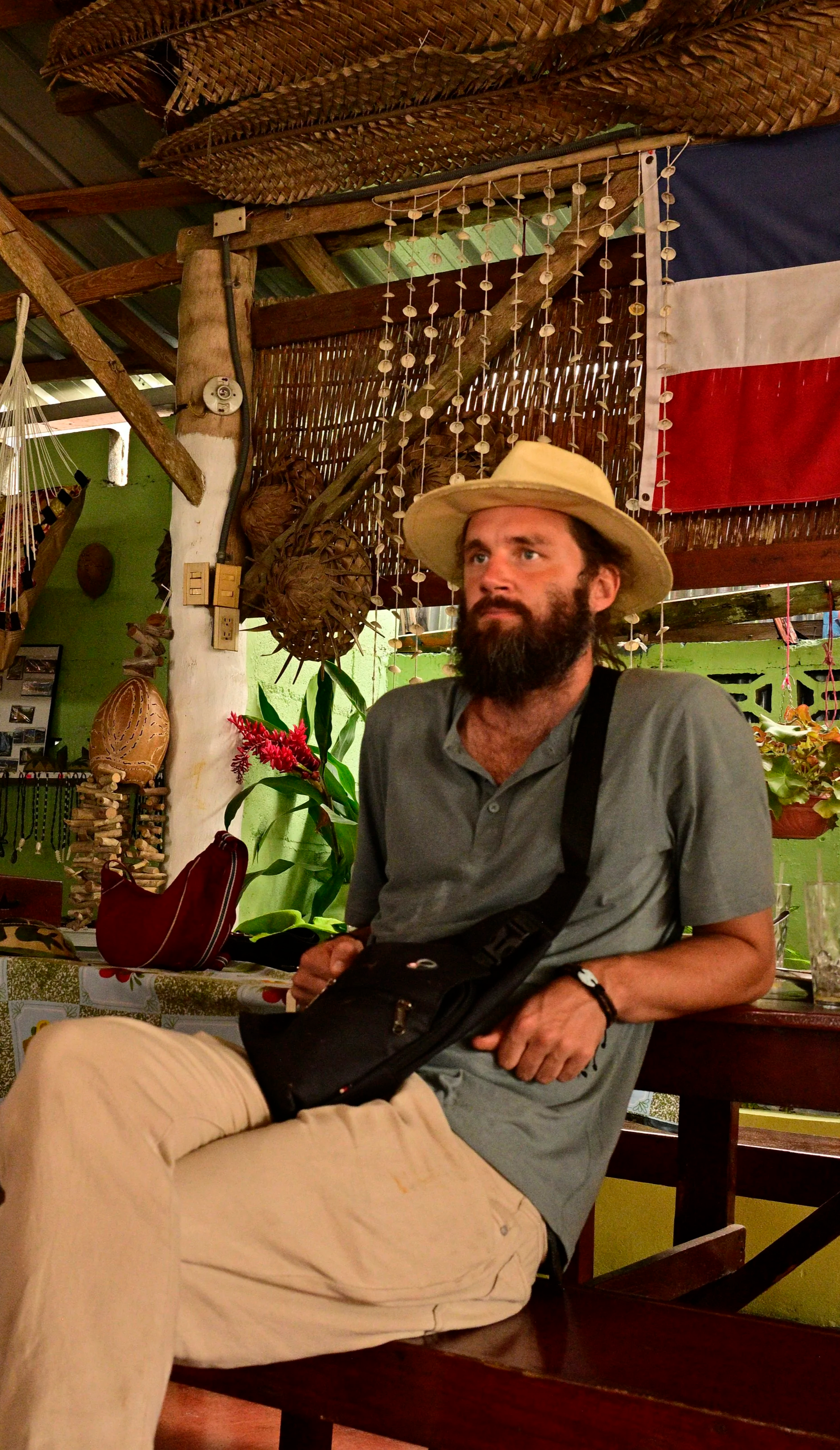 A man with a beard and a mustache wearing a wide-brimmed hat, gray shirt, beige pants, and a black shoulder strap, sitting at a table inside a tropical-themed restaurant with decorative hanging objects, a flag, plants, and woven baskets in the background.