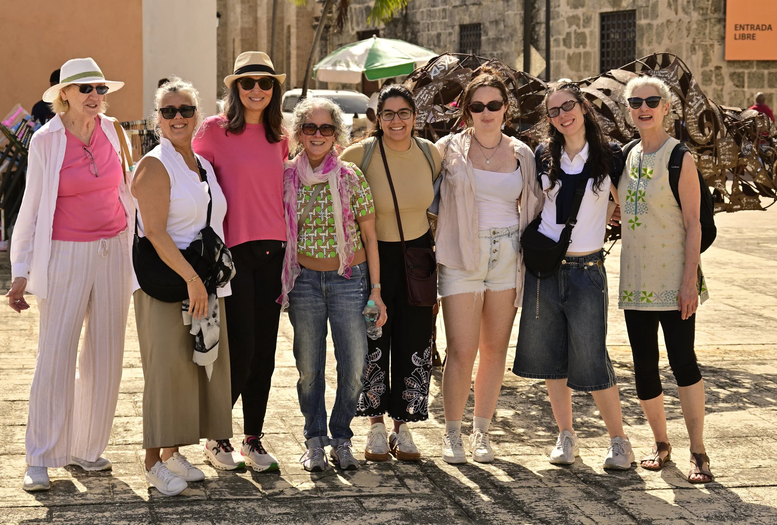 Group of nine women standing together outdoors, smiling, wearing casual summer clothing, sunglasses, and hats, with historical stone buildings and a sculpture in the background.