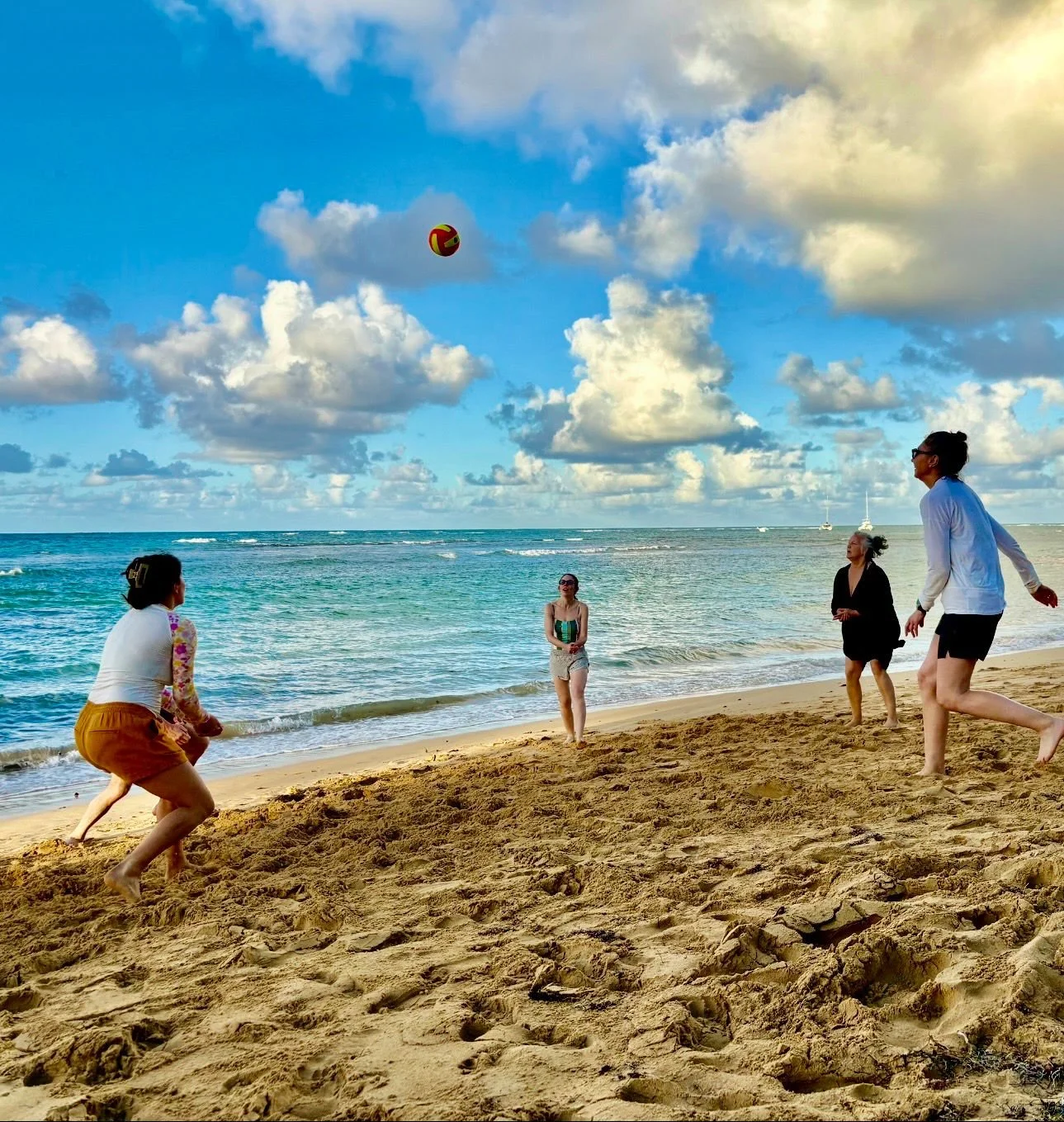 People playing beach volleyball on the sandy shore with ocean and cloudy sky in the background.