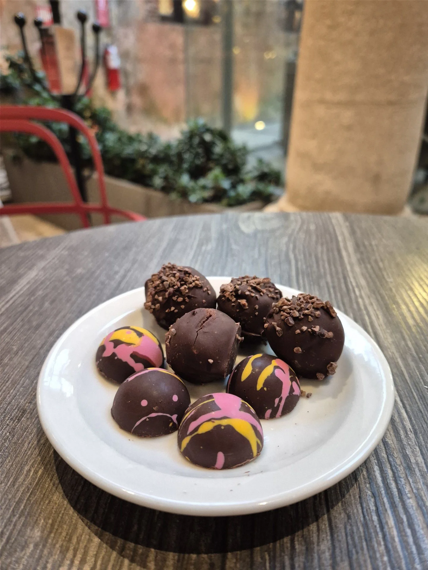 A white plate with assorted truffles covered in dark chocolate, some decorated with pink, yellow, and purple drizzle or sprinkles, on a wooden table.