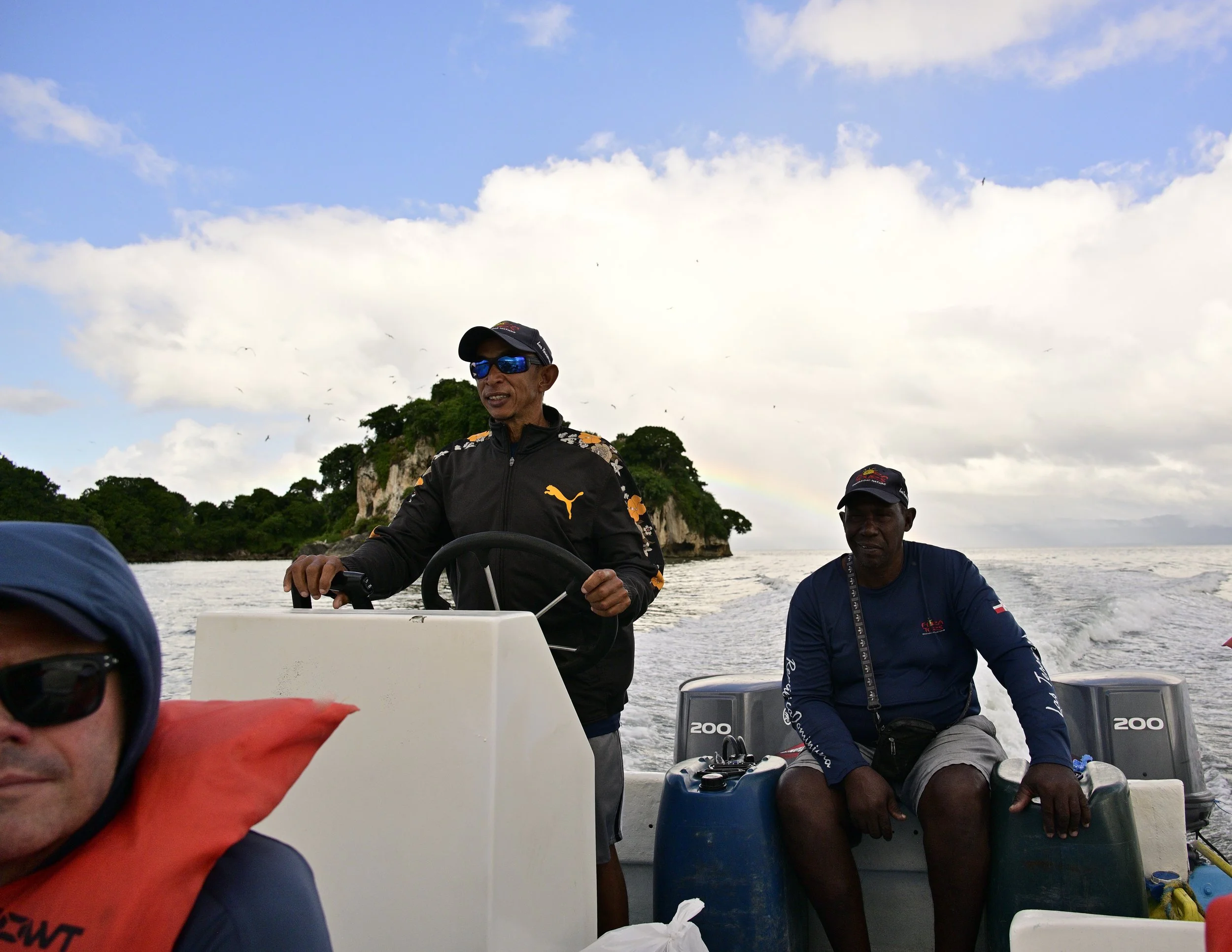 Two men are on a boat with a scenic background of green trees and cliffs, and a cloudy sky. One man is steering the boat, wearing a black cap and sunglasses, and the other man is sitting next to him, wearing a dark blue shirt and a lanyard. Part of a person’s face with sunglasses and a red life vest is visible in the foreground.