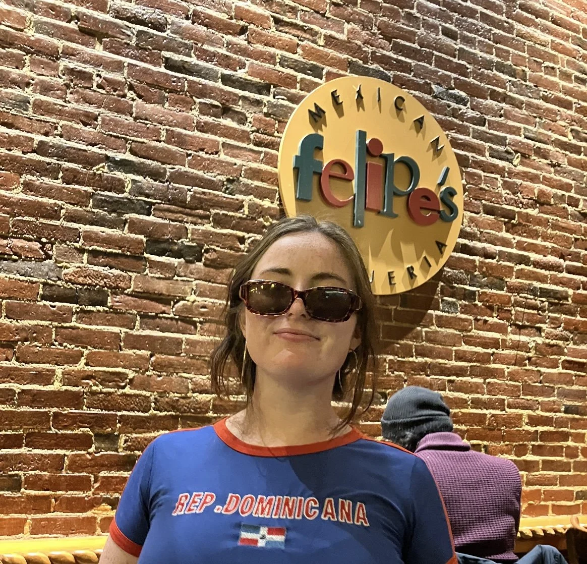 A woman wearing sunglasses and a blue T-shirt with red trim, standing in front of a brick wall with a sign that reads 'Feliz' and 'Mexican' around a yellow circle.