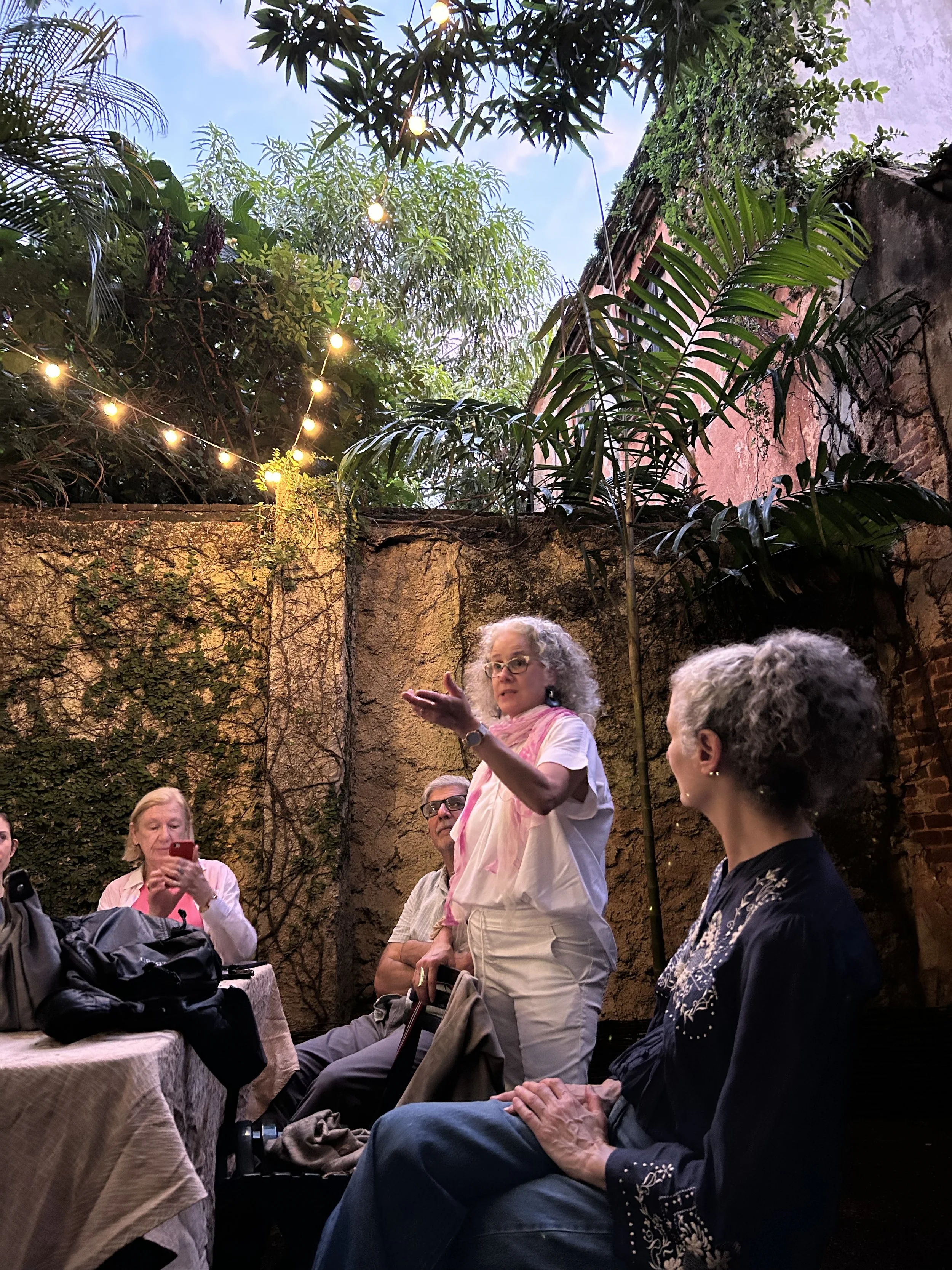 A woman with curly gray hair and glasses gestures while speaking to a group of people seated at a table in a cozy, outdoor courtyard with lush green plants and string lights overhead.