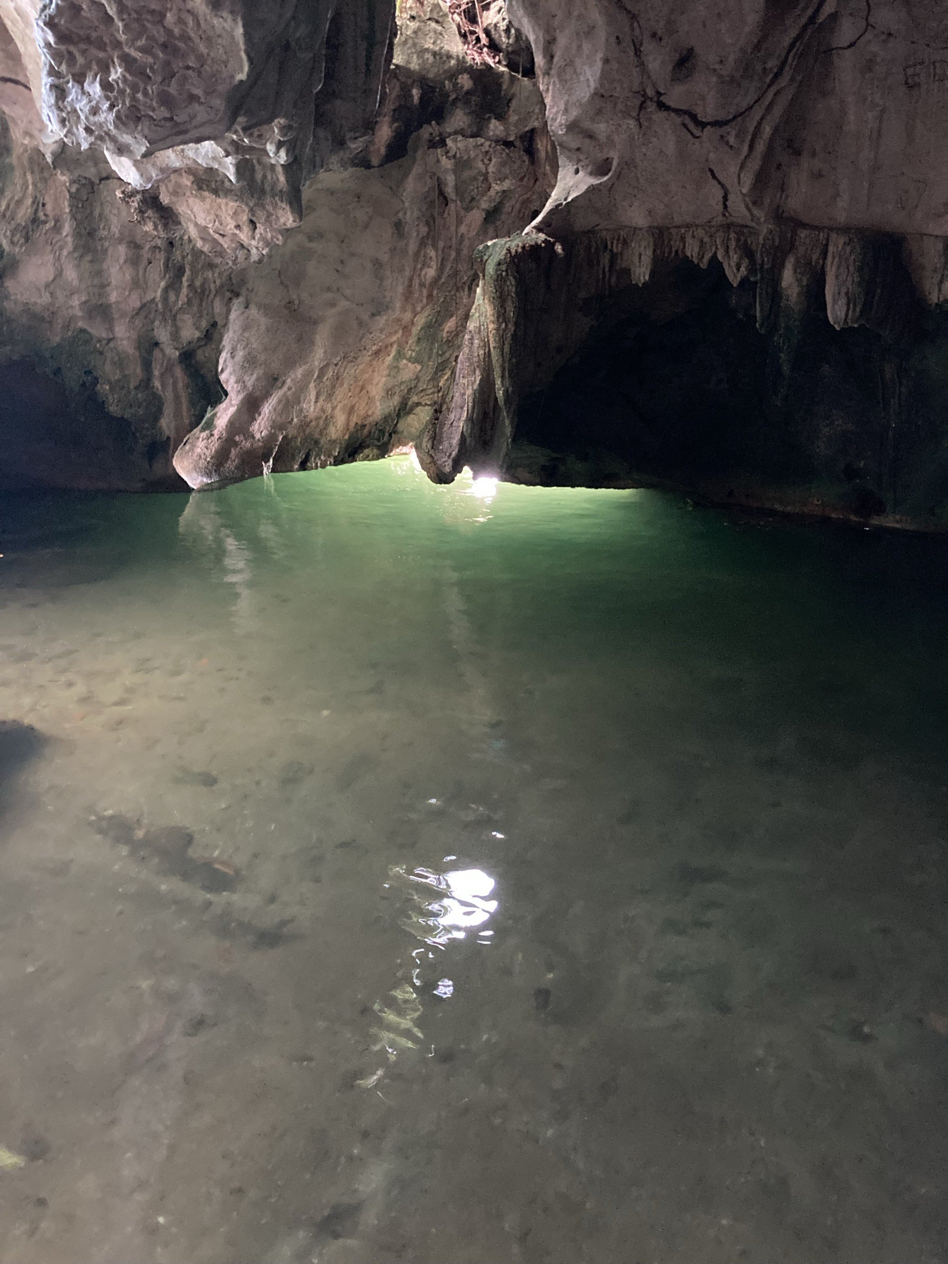 A view inside a dark cave or tunnel with rocky walls and a small opening at the far end letting in light, with water on the ground reflecting the entrance light.
