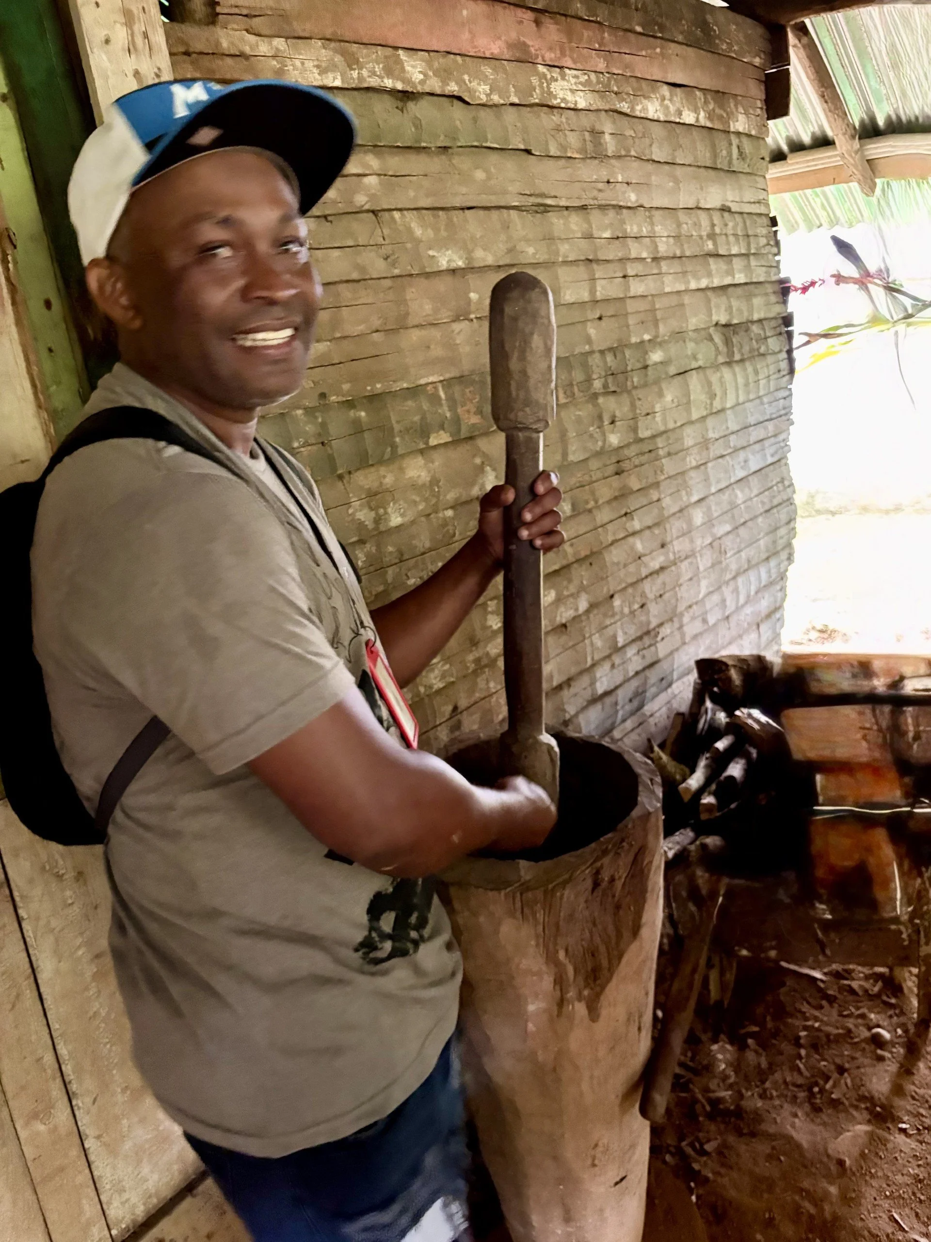 A man wearing a baseball cap and a backpack, smiling, and holding a large wooden pestle inside a rustic wooden building.