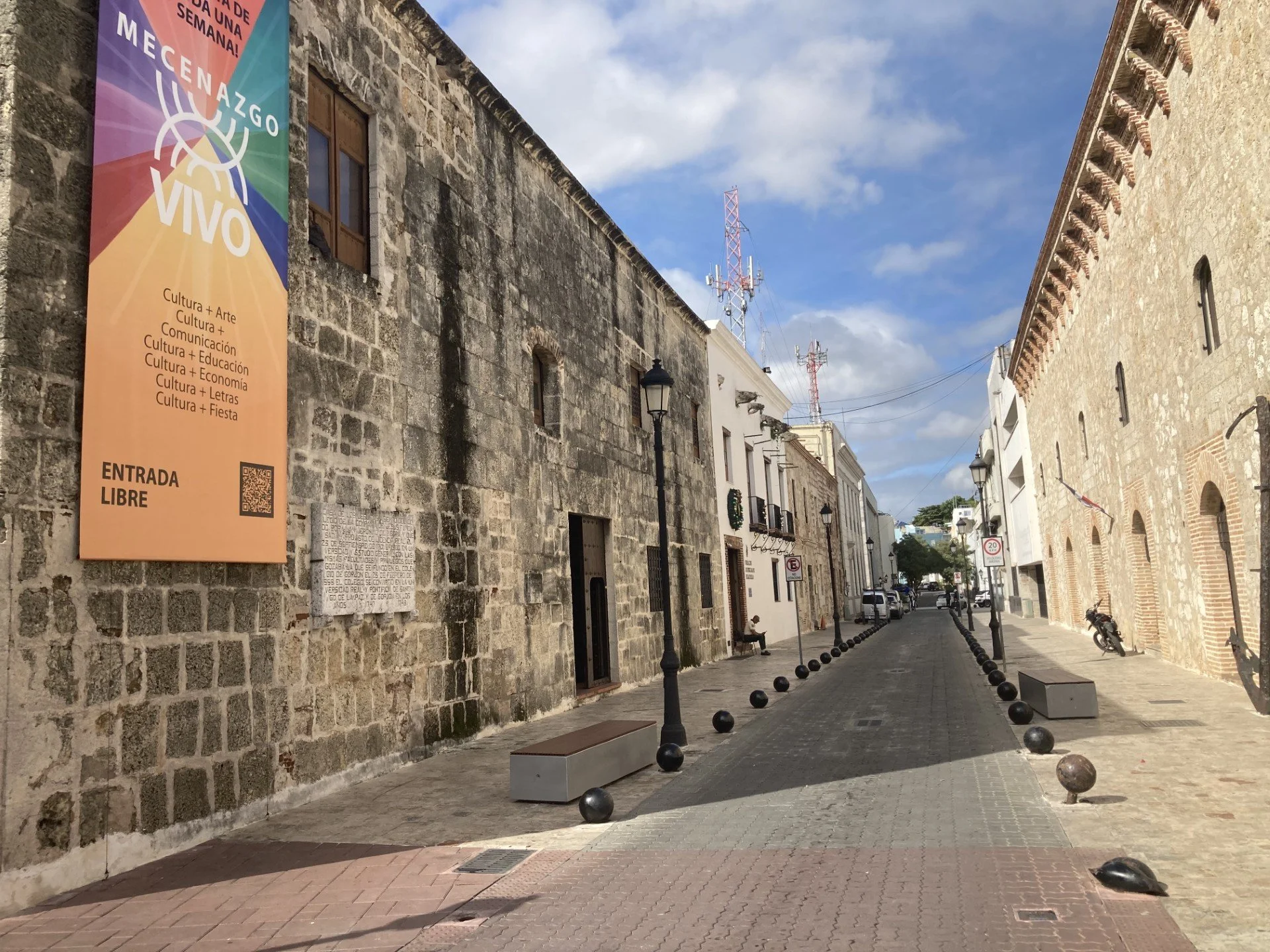 A quiet street with historic stone buildings, black street lamps, and black rounded bollards lining the sidewalk, under a partly cloudy sky.