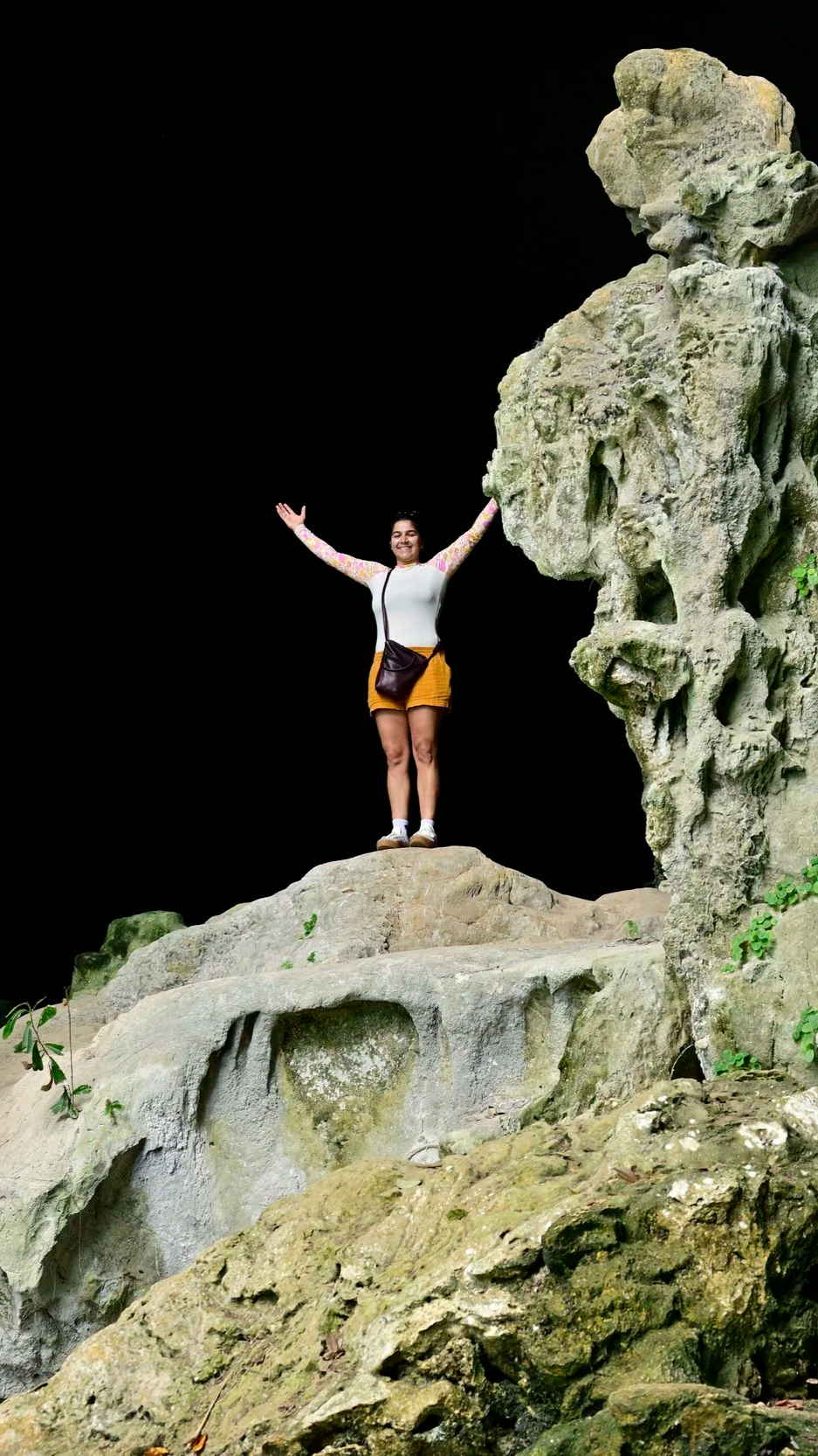 A woman standing on a rock with arms raised, next to an intricate stone formation, against a dark background, smiling.