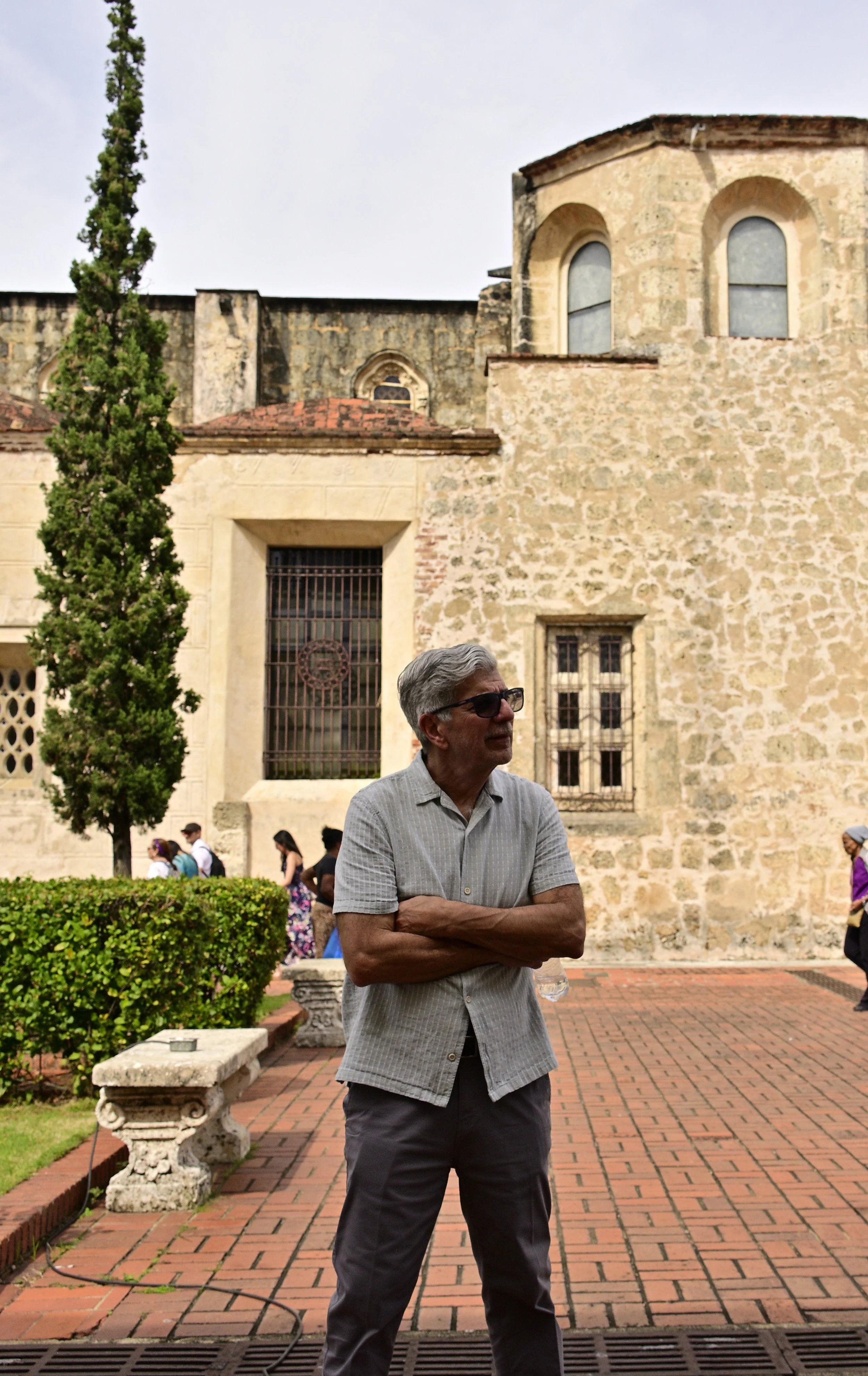 An older man with gray hair and sunglasses standing outdoors near a historic stone building with arched windows, surrounded by a small group of people and greenery.