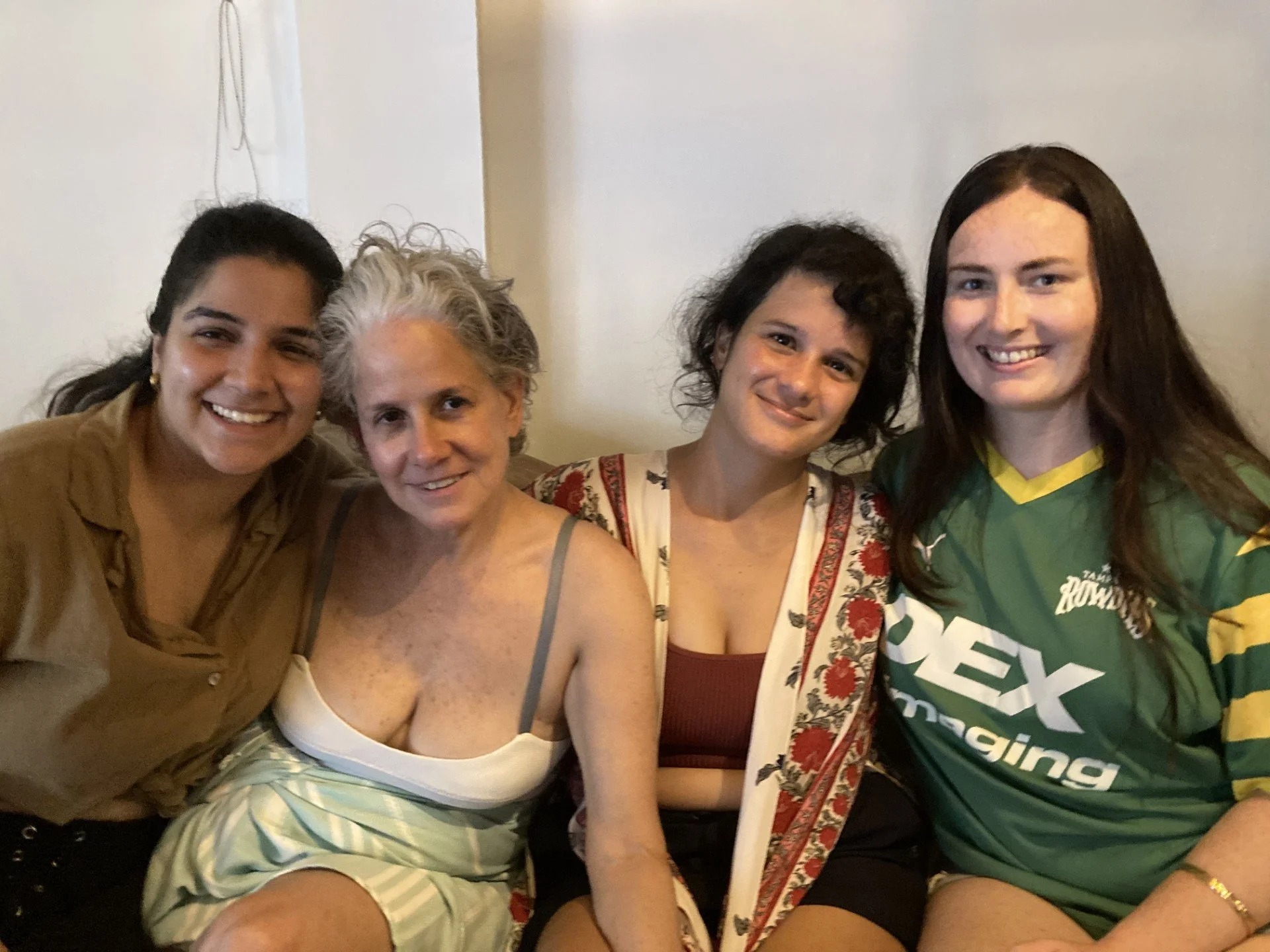 Group of five smiling women sitting close together inside, with plain wall background.