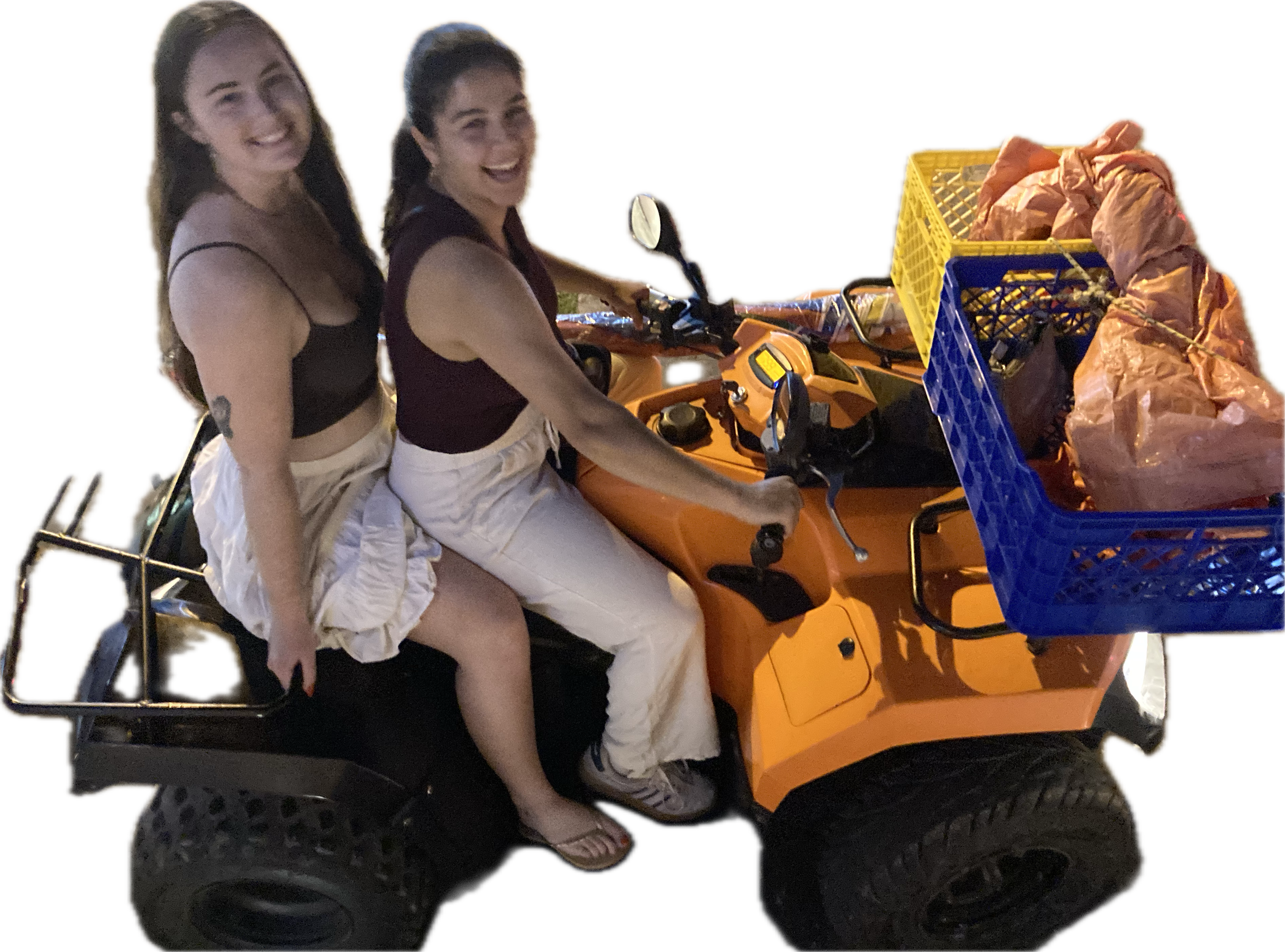 Two women smiling while riding in a small orange utility vehicle, with one woman in the driver seat and the other sitting behind her, and a yellow crate filled with crumpled paper in the front of the vehicle.