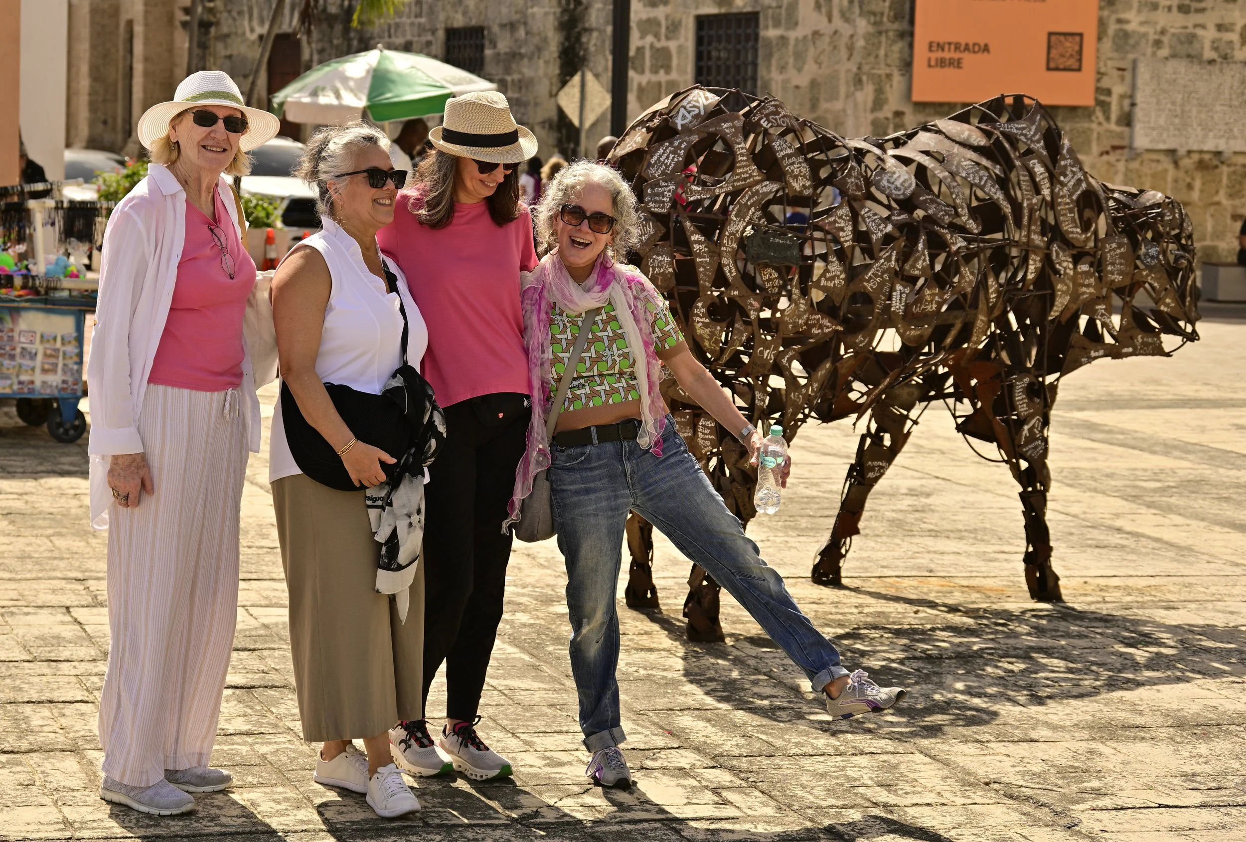 Four women smiling and standing together outdoors, with a horse sculpture covered in written messages behind them.