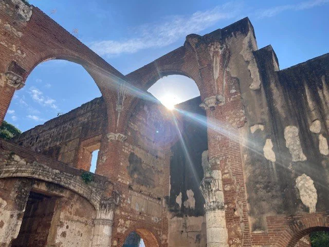 Photo of the ruins of an old brick building with arches, with the sun shining through a circular opening in the structure, set against a blue sky with some clouds.
