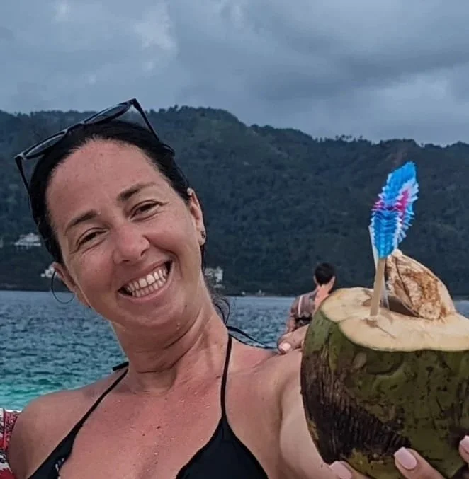 Woman smiling and holding a coconut with a small umbrella in it on a boat, with a body of water and forested hills in the background.