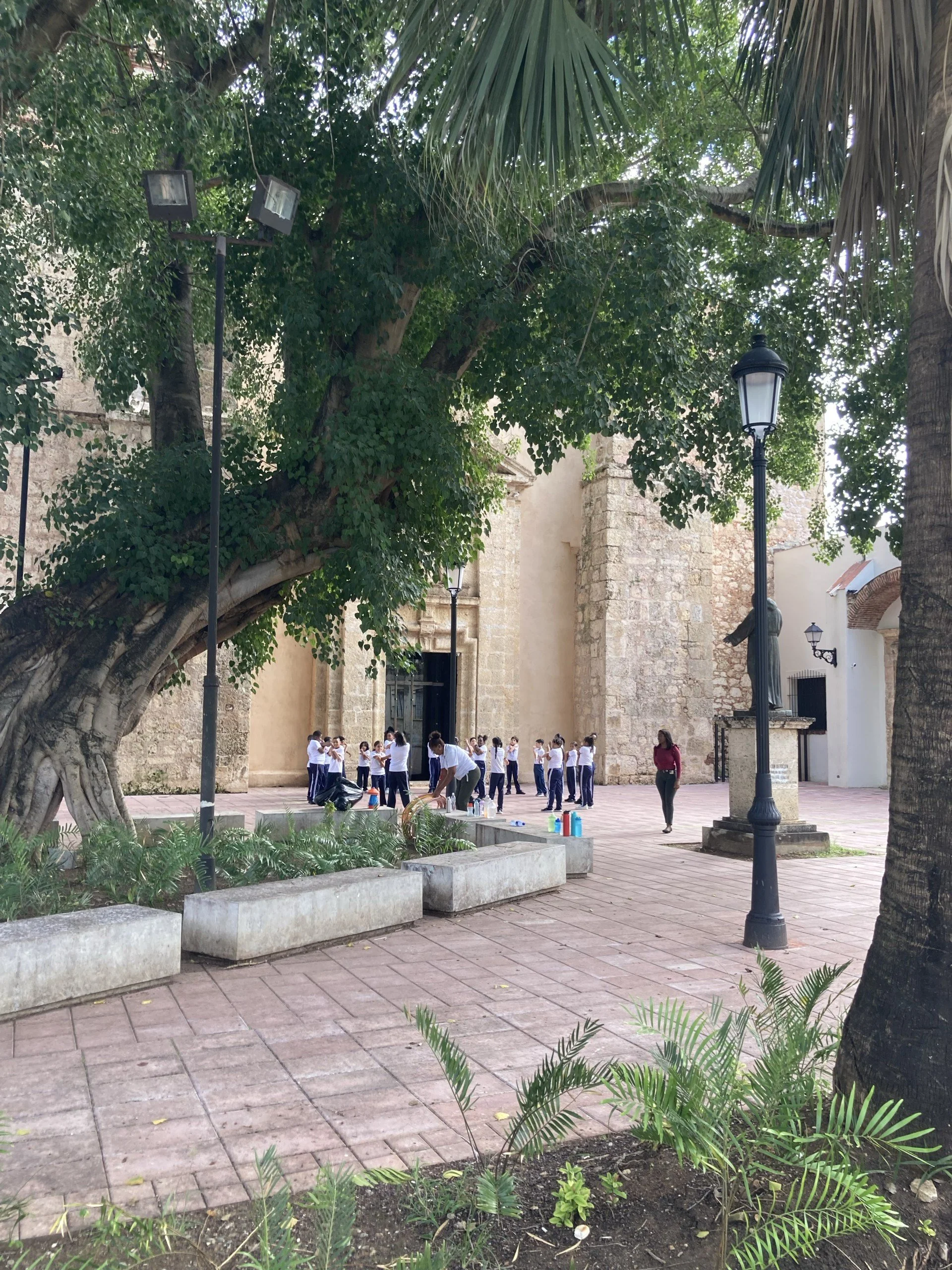 Children in school uniforms stand in a courtyard with large trees and historic stone building in the background, some adults are nearby.