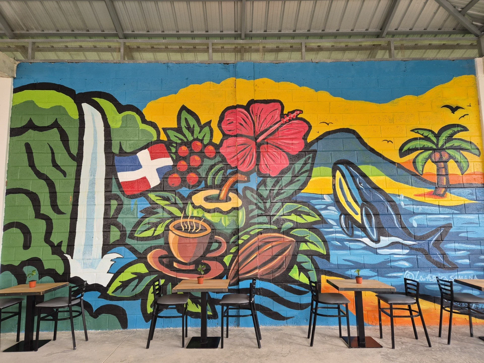 Colorful mural of a tropical scene with a large red hibiscus flower, a toucan, palm trees, and mountains in the background, painted on a brick wall with tables and chairs in front.