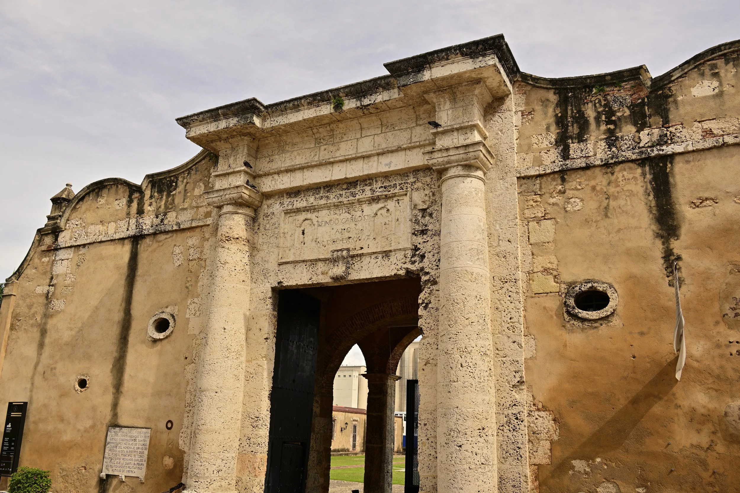 Ancient stone archway with columns, weathered walls, and a plaque, likely part of historic ruins or a heritage site, under a cloudy sky.