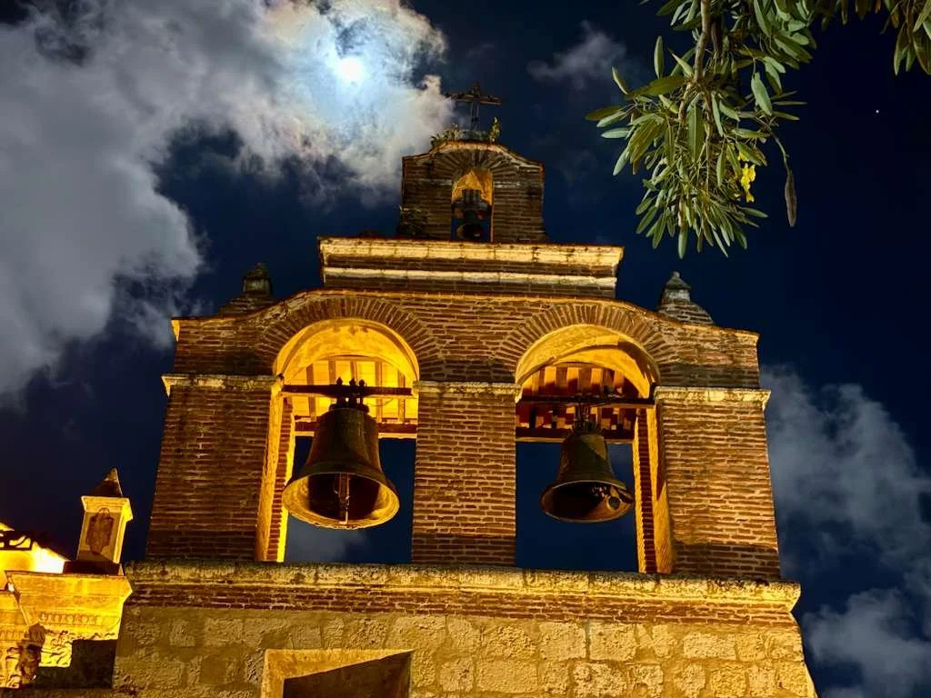 Night view of a historic brick church bell tower illuminated by warm yellow lighting, with the moon and clouds in the sky and tree branches partially visible.