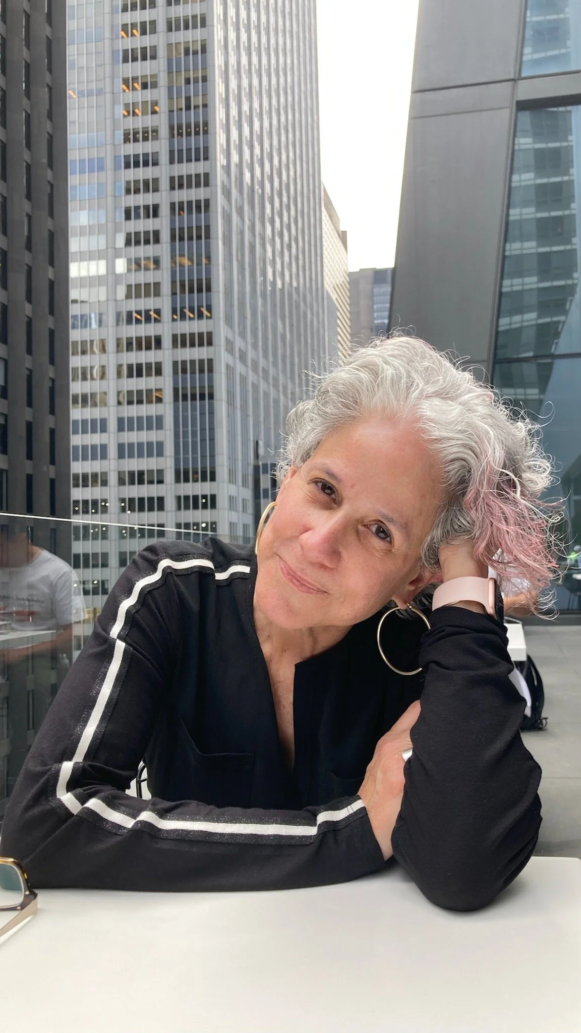 A woman with curly gray hair and large hoop earrings sitting at a table in an urban outdoor setting with tall glass skyscrapers in the background, wearing a black tracksuit with white stripes.