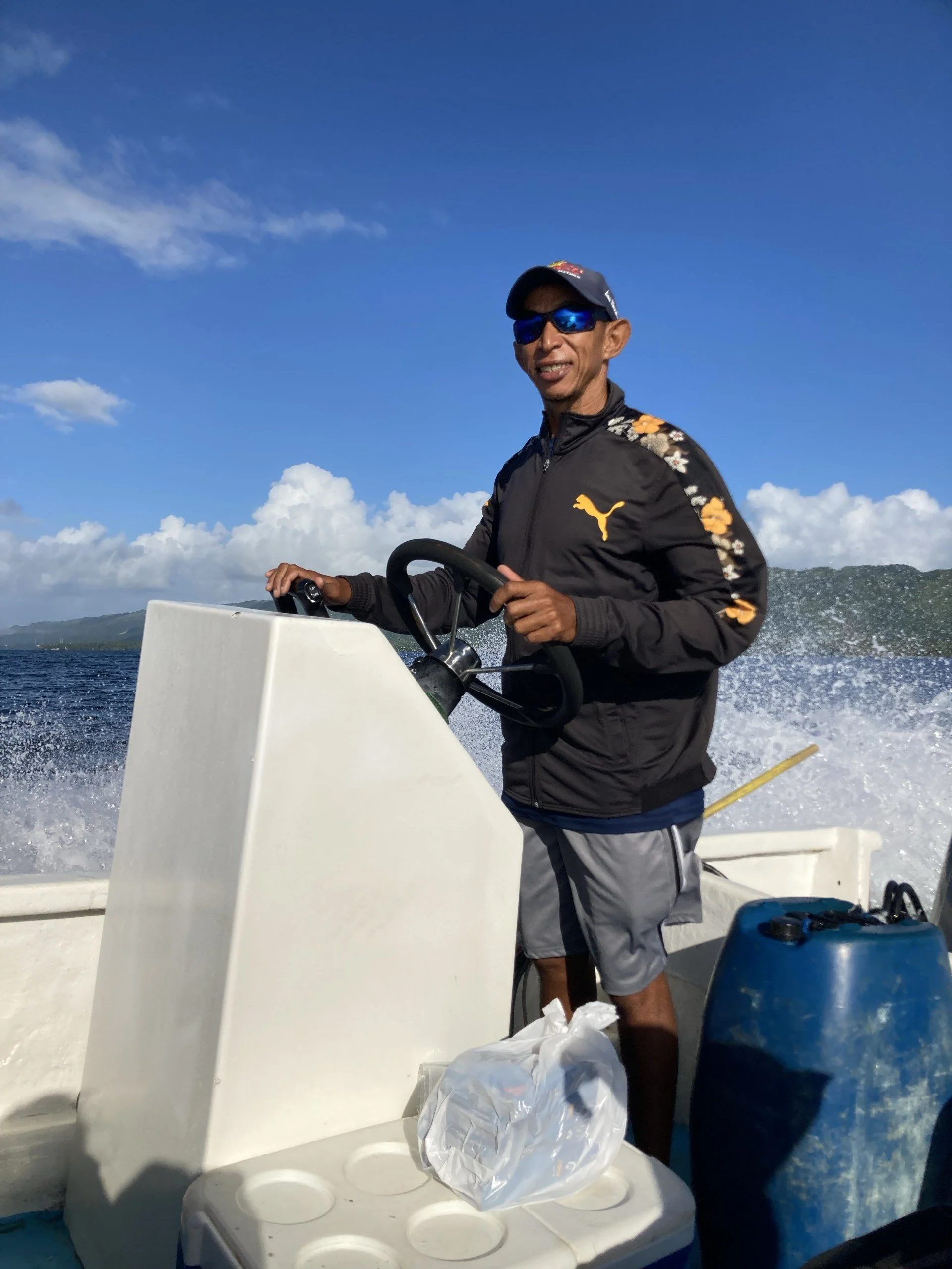 A man steering a boat on open water with a blue sky and trees in the background. He is wearing sunglasses, a cap, and a jacket with a Puma logo.