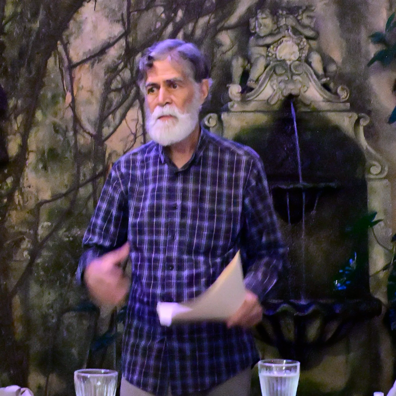 An elderly man with white hair and beard, wearing a checkered shirt, is speaking or giving a presentation in a rustic setting. There are two glasses of water on the table in front of him.
