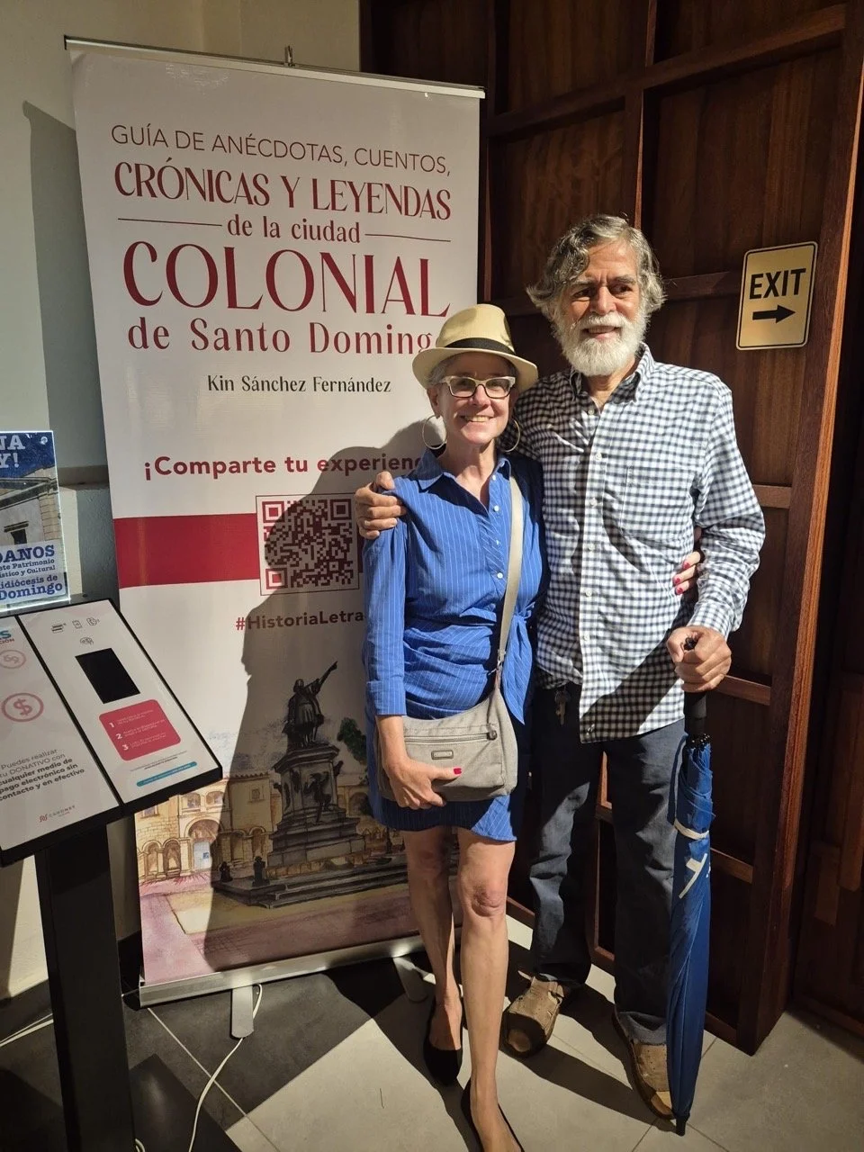 A smiling woman and man standing together indoors, with the woman wearing a tan hat, glasses, a blue dress, and carrying a beige purse, and the man holding a blue umbrella, both smiling at the camera. Behind them is a sign in Spanish about the colonial history of Santo Domingo, Dominican Republic, and a wooden wall with an 'EXIT' sign.