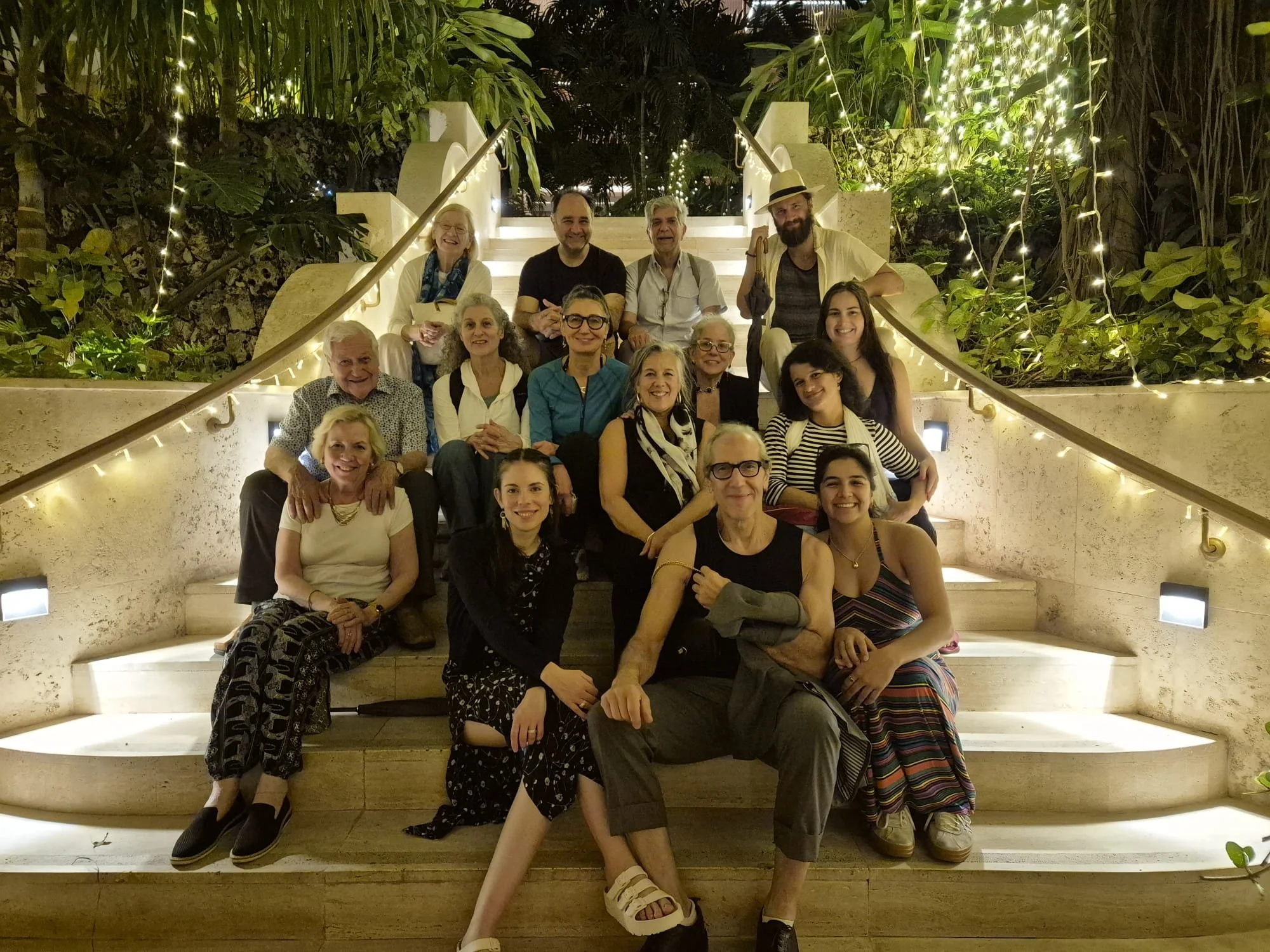A group of 17 people, including older adults and younger adults, gathered on a staircase at night, surrounded by lush greenery and decorated with string lights, smiling and posing for the photo.