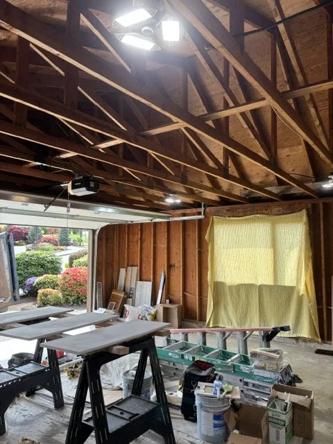 Interior of a garage or workshop with wooden ceiling trusses, a partially open garage door revealing bushes and flowers outside, and various tools and construction materials scattered inside.