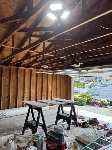 An unfinished garage with exposed wooden beams, construction tools, workbenches, and a view of a driveway and neighborhood outside.