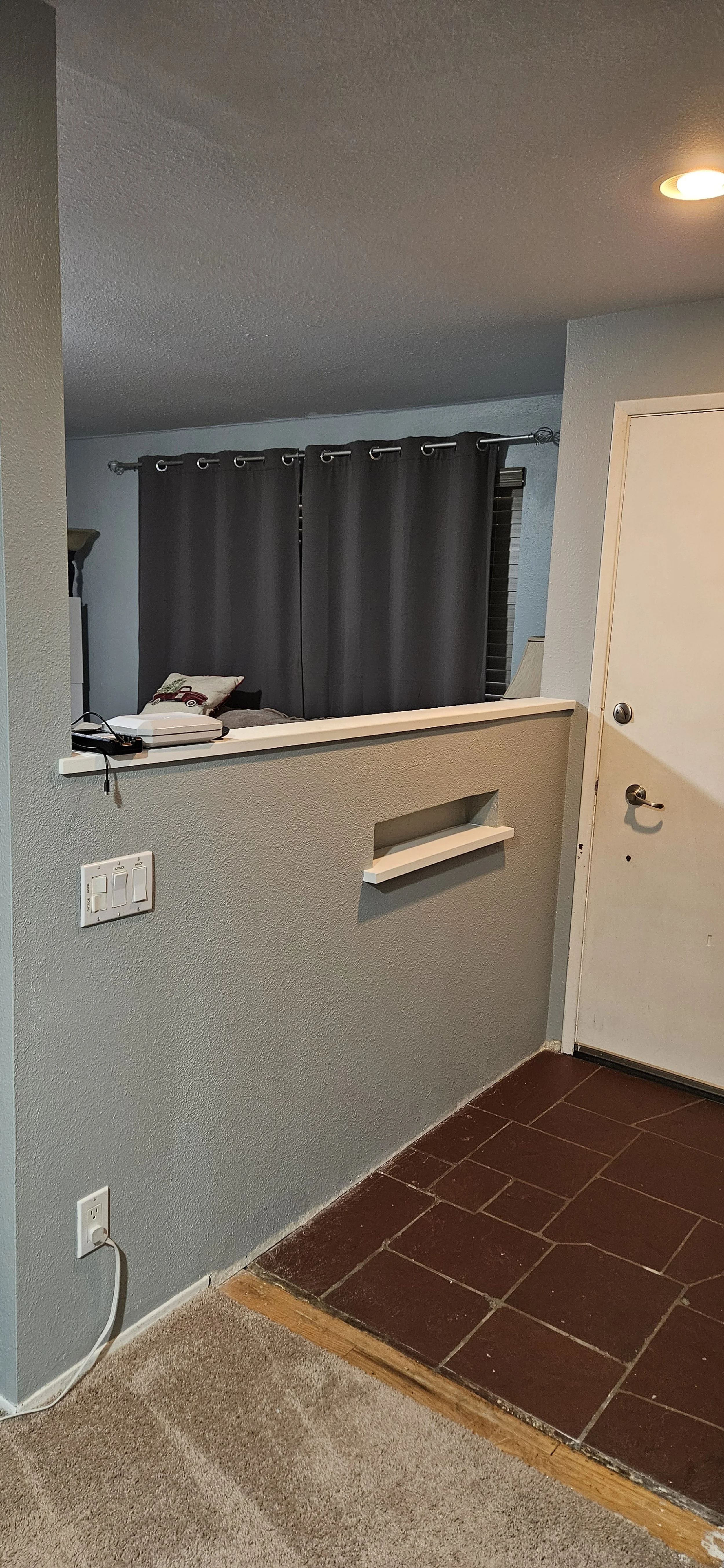 Interior view of a home showing a hallway with a small step down to a tiled floor, a door painted white, a wall with light switches and an outlet, and a partial view of a living room with dark curtains and a bed.