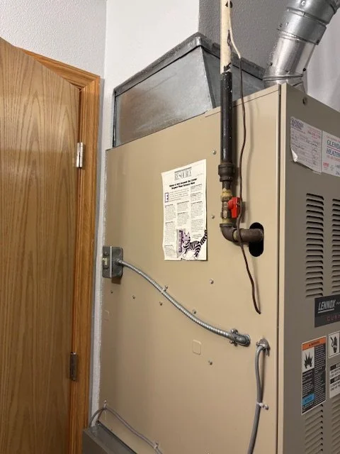A beige furnace or HVAC unit installed indoors next to a wooden door, with various pipes, wires, and a venting duct connected to it.