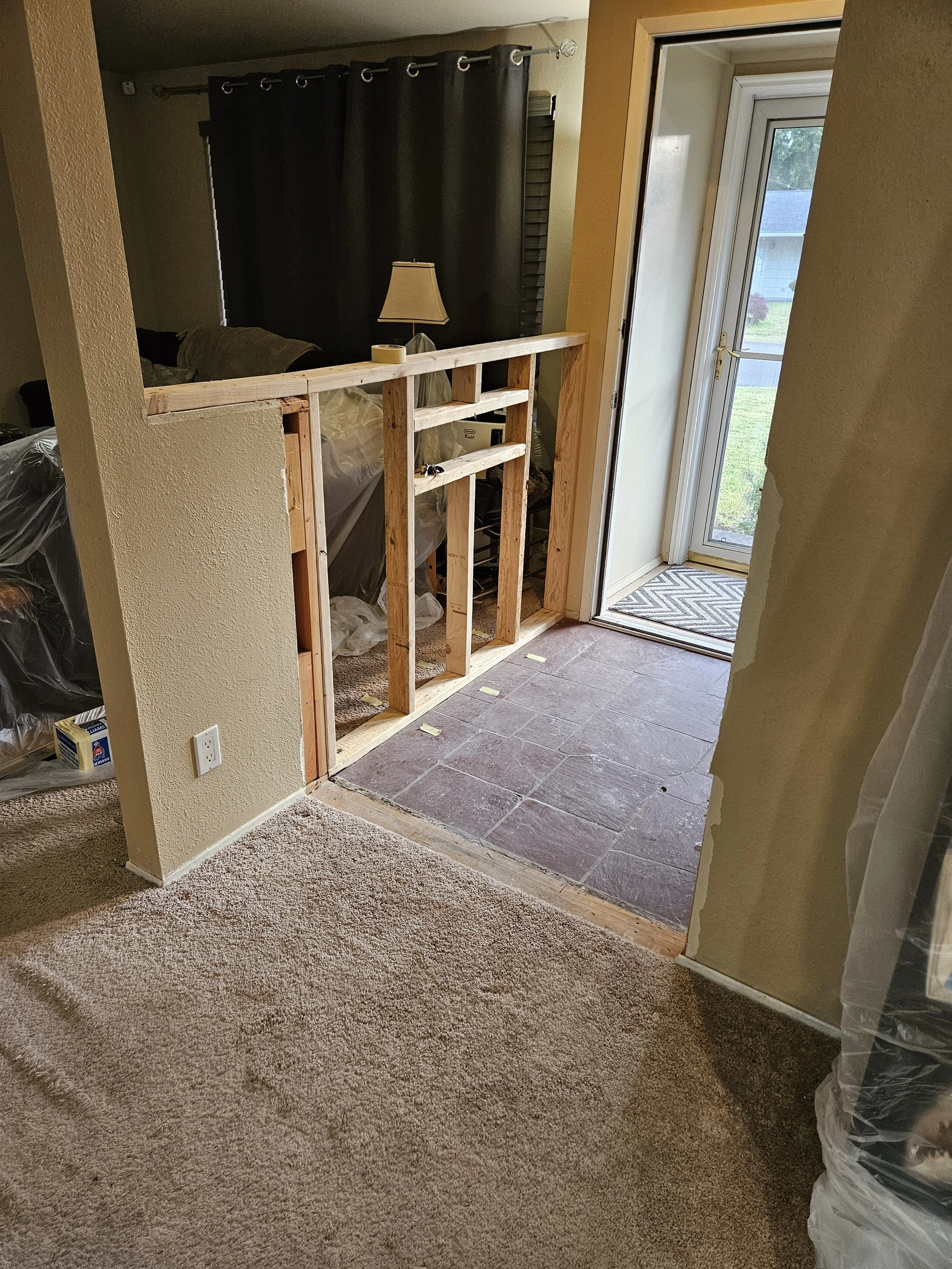 Interior view of a room under renovation with a partially built wooden frame around an opening, leading to an outdoor area with a door and window. Carpeted floor in the foreground, tile floor beyond the frame, and dark curtains on a window or doorway in the background.