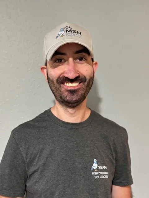 A smiling man wearing a dark gray t-shirt and a white cap that has a logo with a mountain and the letters "MSH." The t-shirt has a logo with a duck and the text "SEAN MSH DRYWALL SOLUTIONS." He is standing against a plain light-colored wall.