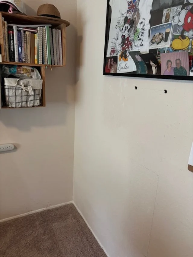 Corner of a room with a wall-mounted bookshelf holding books and a wire basket, and a bulletin board with various pictures and artwork.