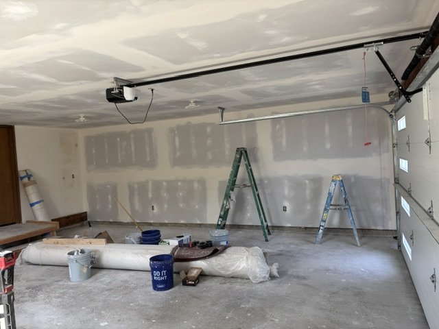 Garage with drywall in progress, two metal ladders, construction tools, and supplies scattered on the floor.