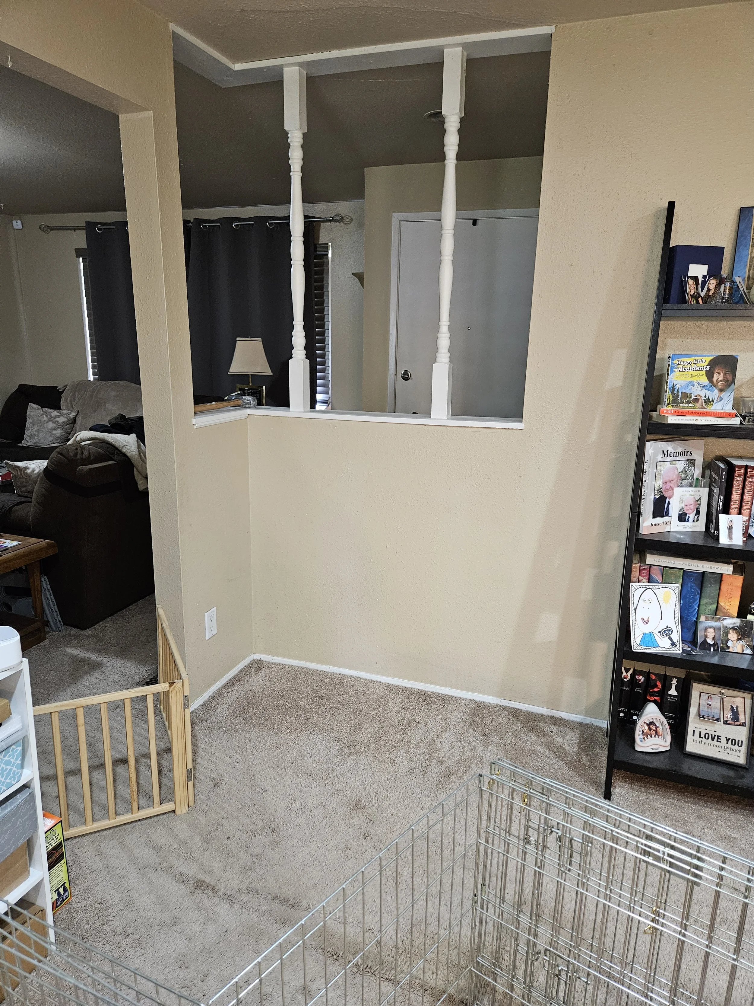 Living room with beige walls, a brown sofa, a bookshelf with books and framed photos, a pet gate, and a metal pet playpen, viewed from a corner near the entrance.