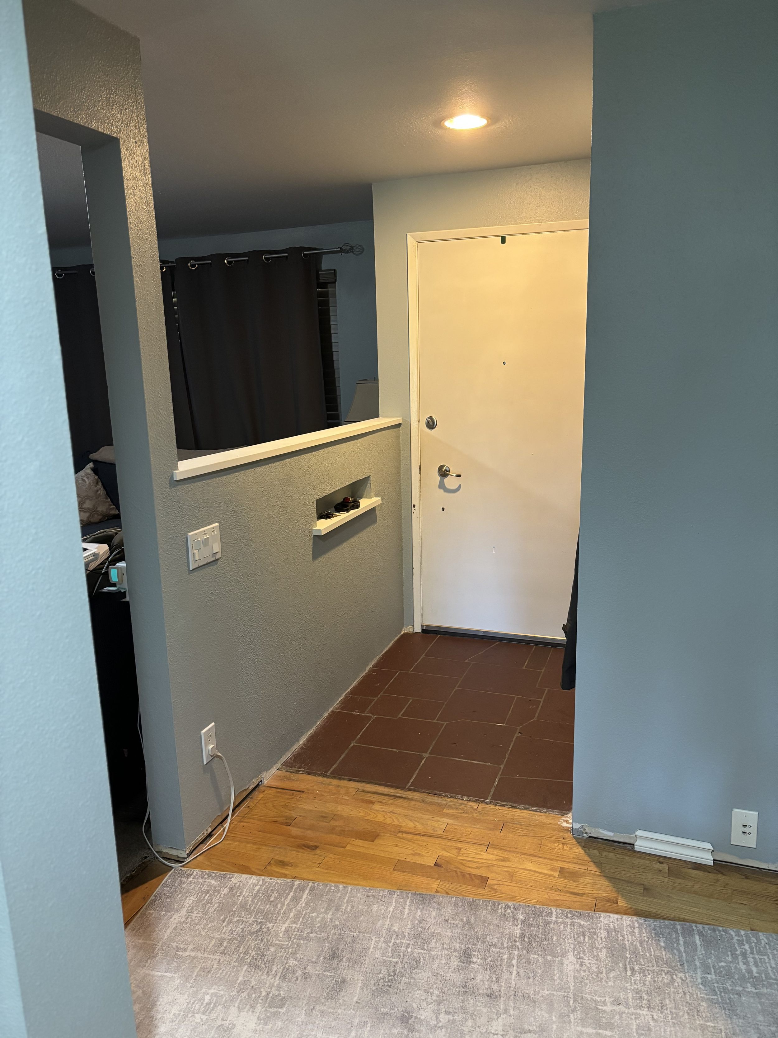 Interior entryway with white door, brown tiled flooring, light blue walls, and a partial view of connecting room with light brown wood flooring and gray area rug.