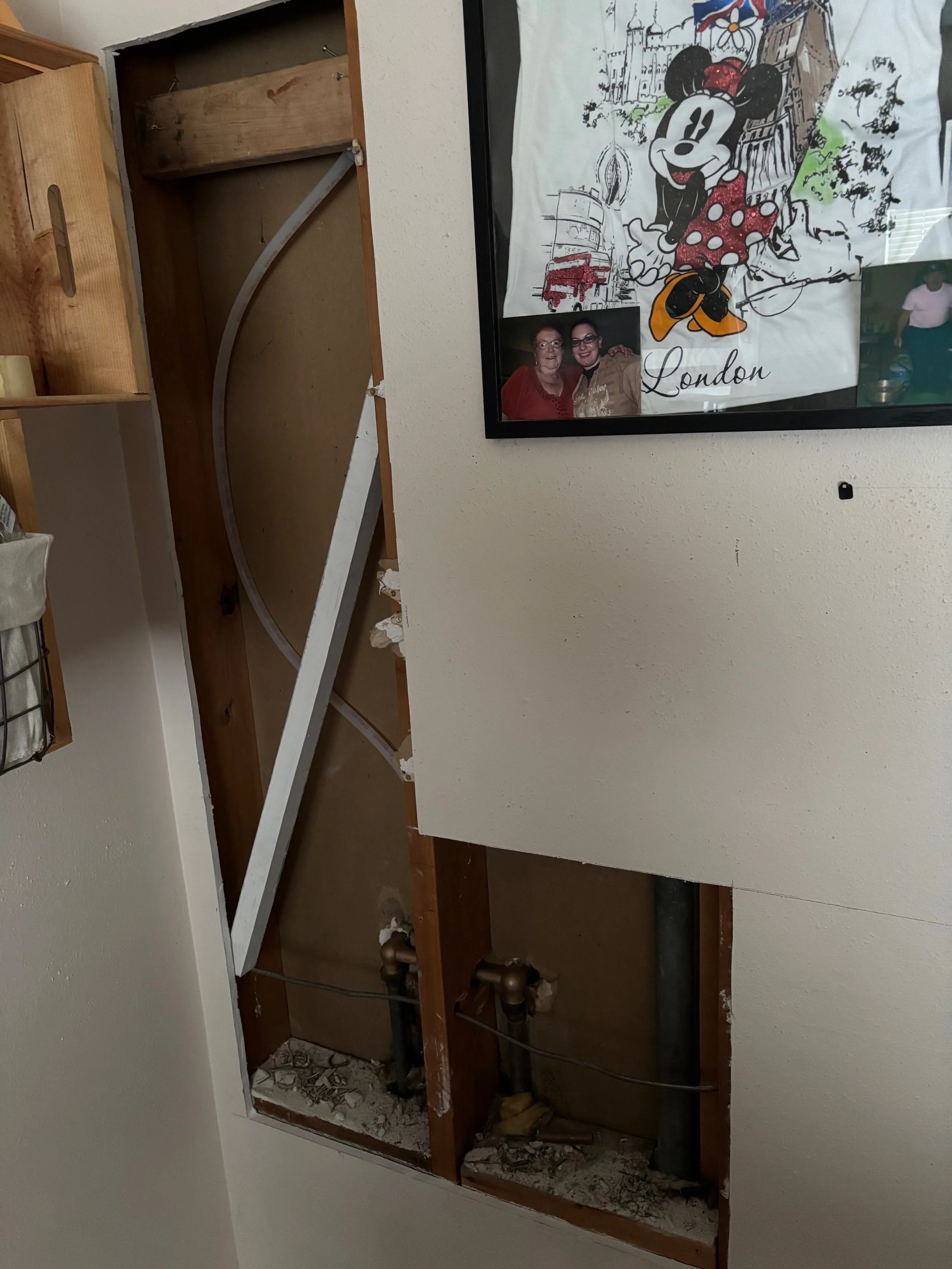 Partially removed wall revealing plumbing pipes, with a framed photo collage featuring a Mickey Mouse drawing, two women, and a person.