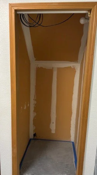 A small, unfinished closet or storage space with drywall and drywall mud, with exposed wiring at the top and a light bulb in the upper right corner.
