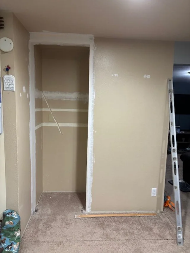 A small empty closet frame under construction with a partially painted wall inside. A level tool is on the floor next to it, and a ladder is leaning against the wall on the right.