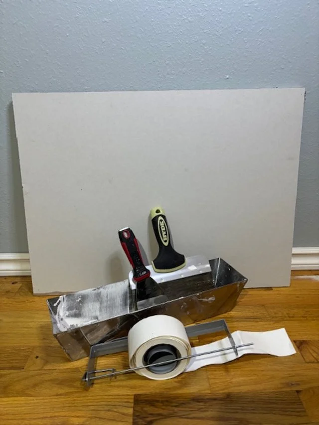 A construction workspace with tools including a metal drywall mud pan, a utility knife, a clean putty knife, a roll of painter's tape, and a sheet of drywall leaning against the wall on a hardwood floor.