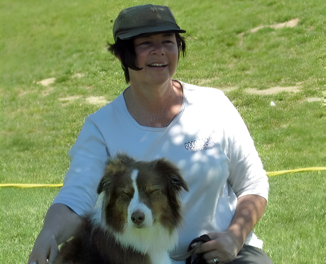 A woman wearing a black cap and white T-shirt smiling with her eyes closed while holding an Australian Shepherd dog with closed eyes outdoors on a sunny day.