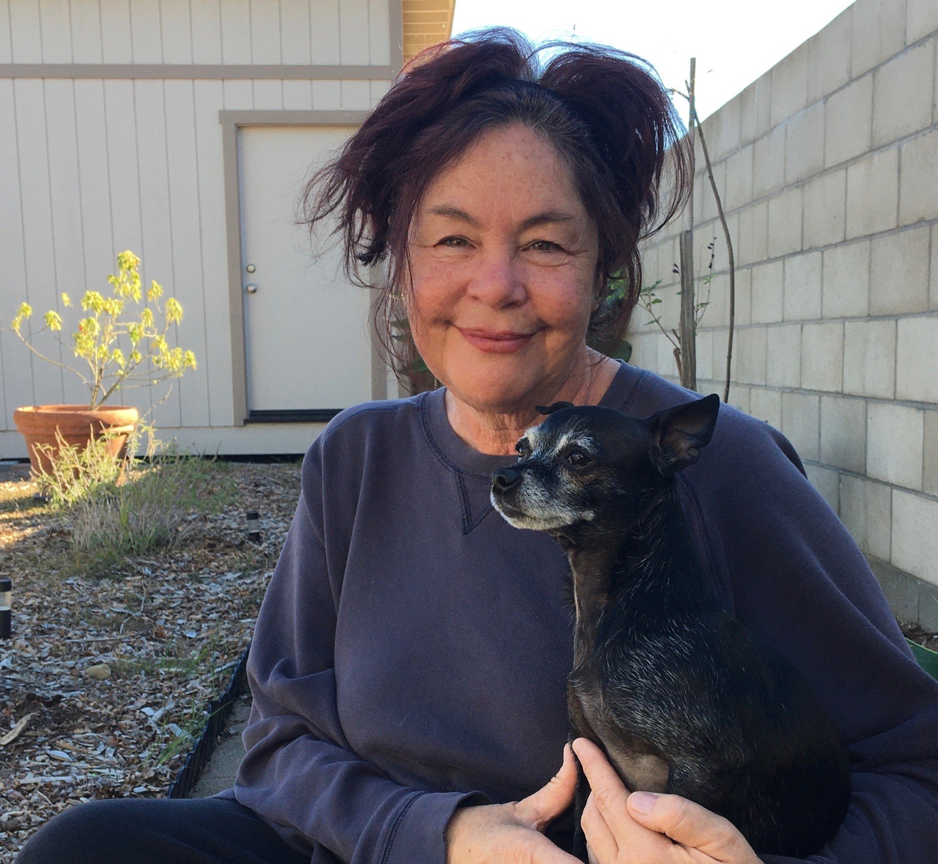 An older woman with short, tousled dark hair smiling while holding a small black dog with gray markings in her arms outdoors. There is a white fence, a potted plant, and a brick wall in the background.