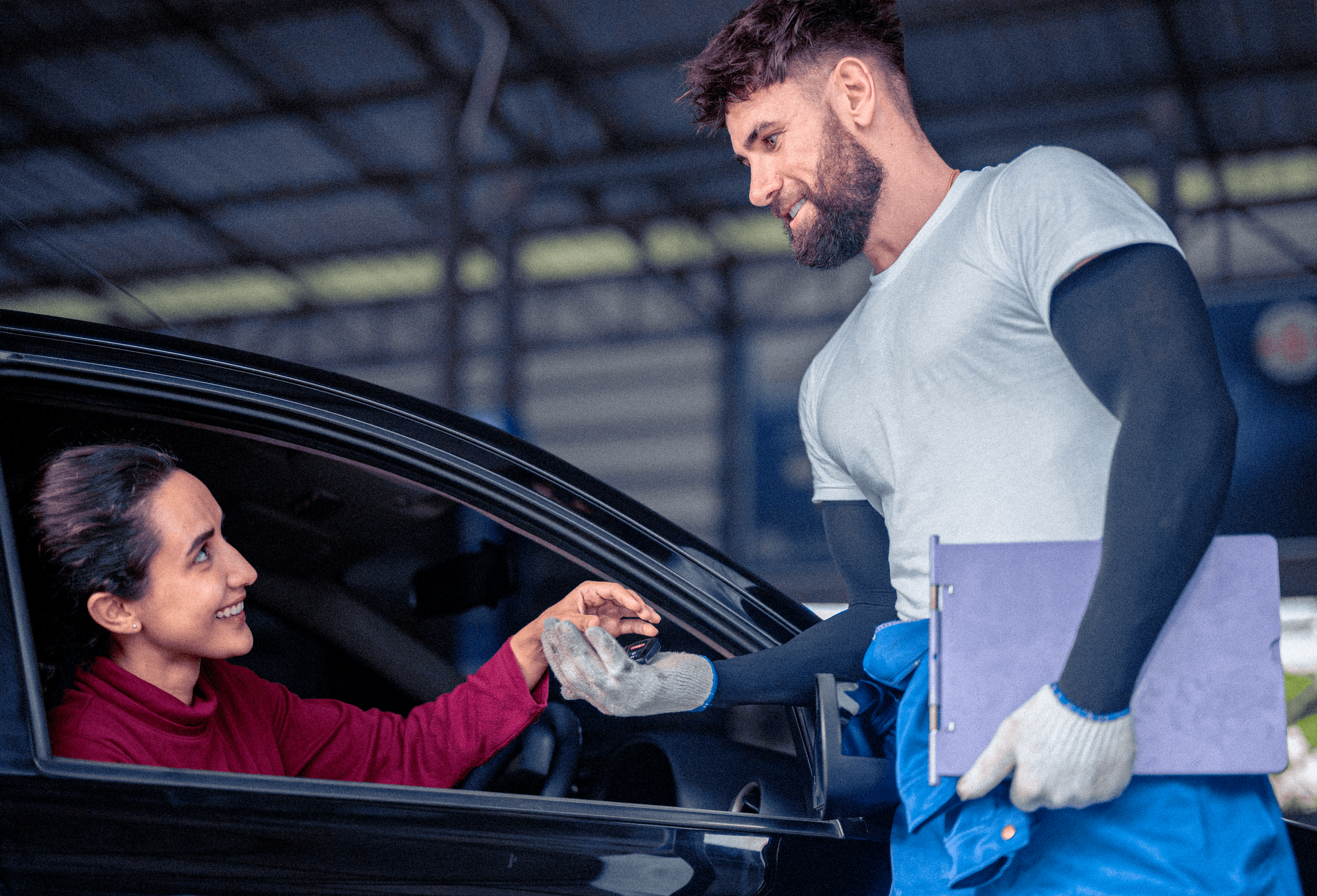 A woman sitting in a car, smiling and holding hands with a man standing outside the car, who is wearing work gloves and holding a clipboard, inside a garage or workshop.