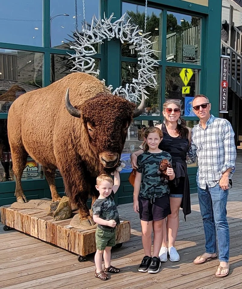 A family of four standing next to a large bison statue with antlers in front of a store with glass windows and a yellow pedestrian sign. The family includes two children and two adults, all smiling in Jackson, Wyoming