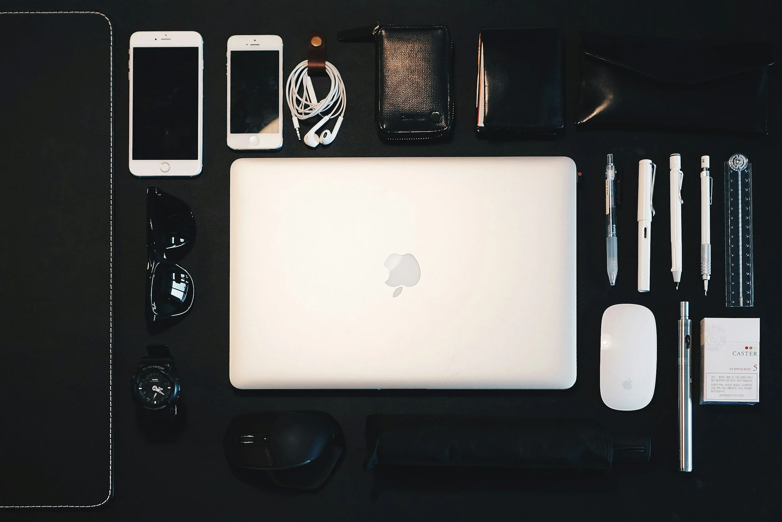 Apple MacBook laptop surrounded by two smartphones, earphones, sunglasses, watch, and various stationery items on a black desk.
