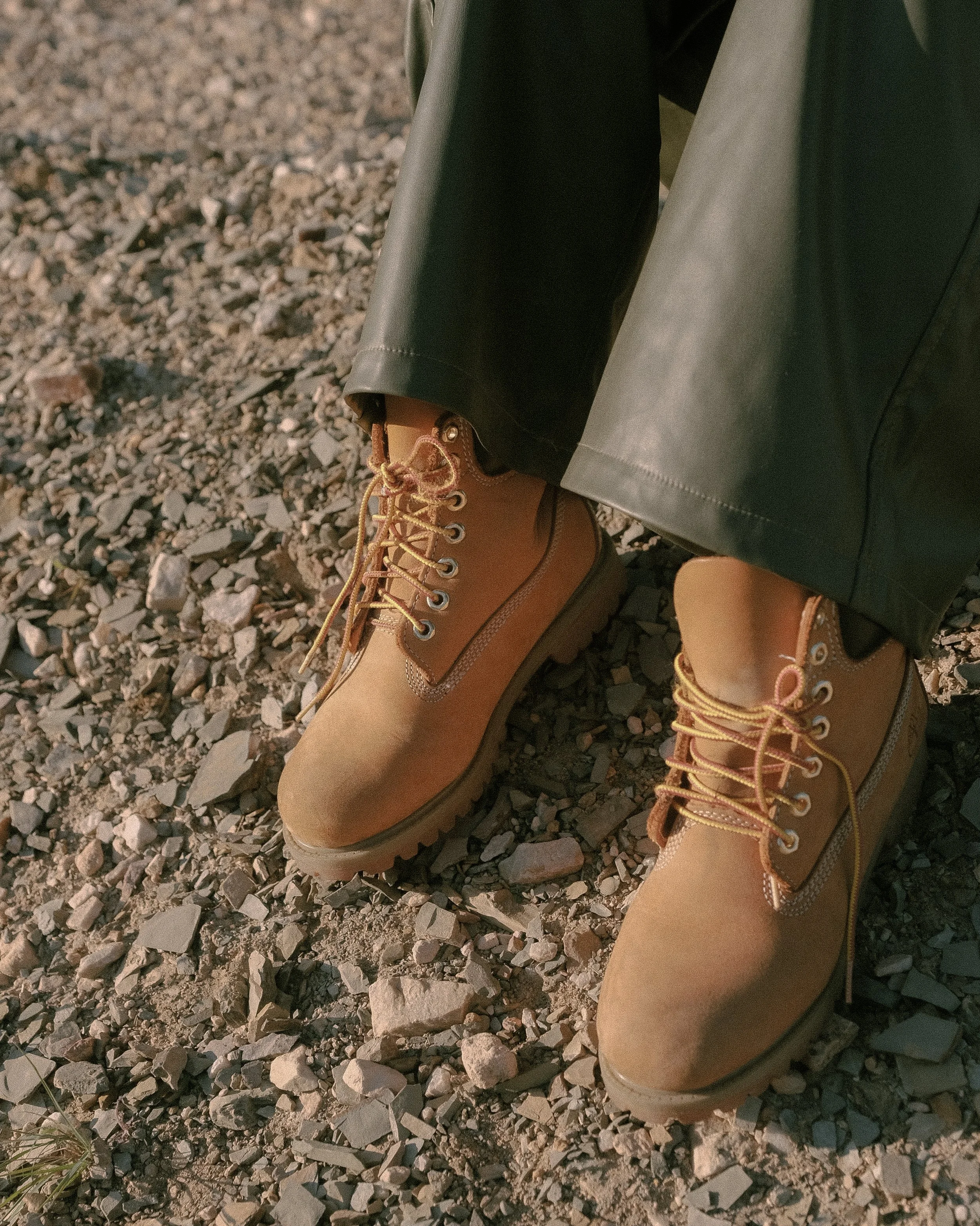 Close-up of a person wearing tan leather boots and dark green pants, standing on a gravel surface.