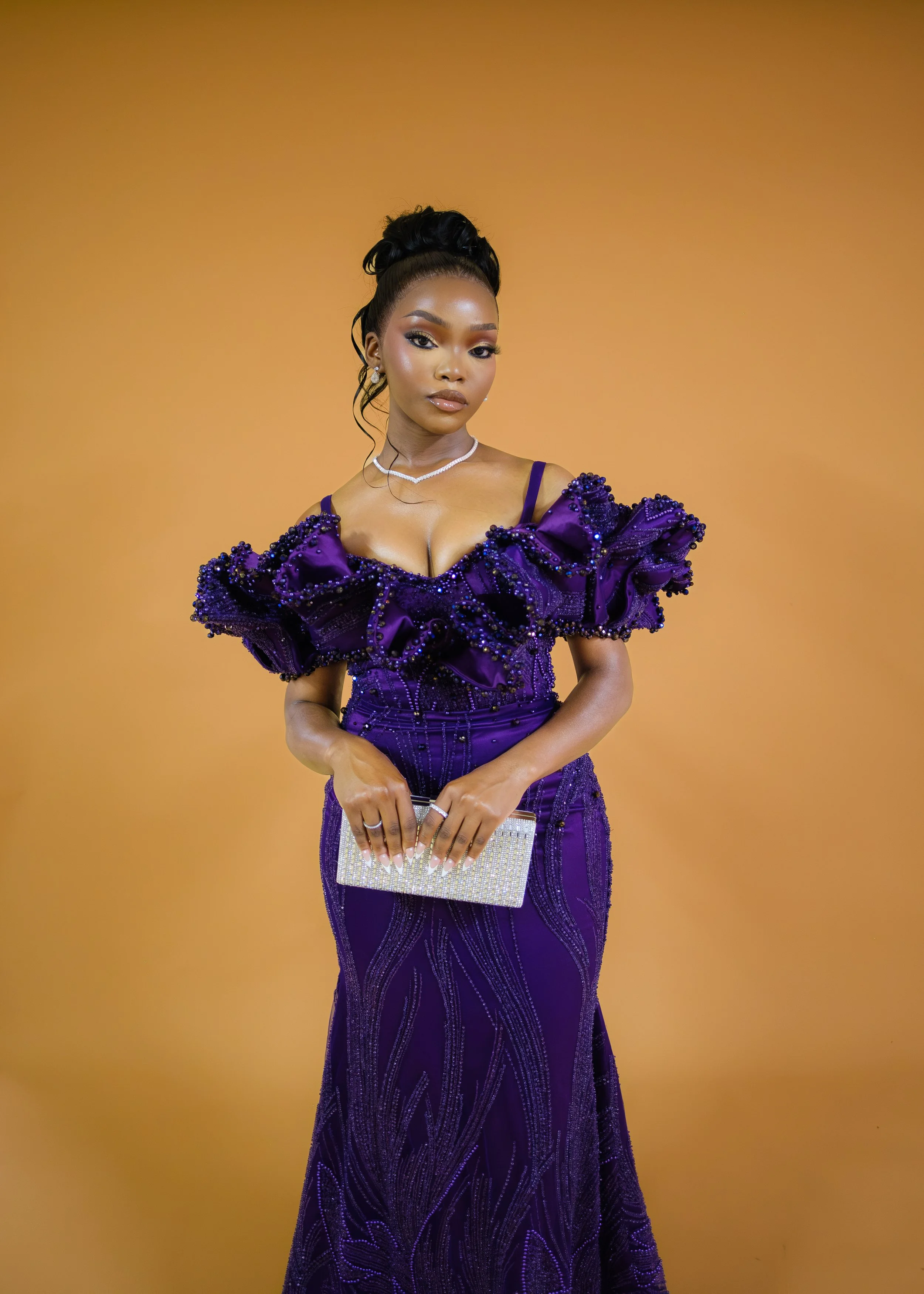 A woman in a purple gown decorated with beads and ruffles, holding a silver clutch, standing against a plain orange background.