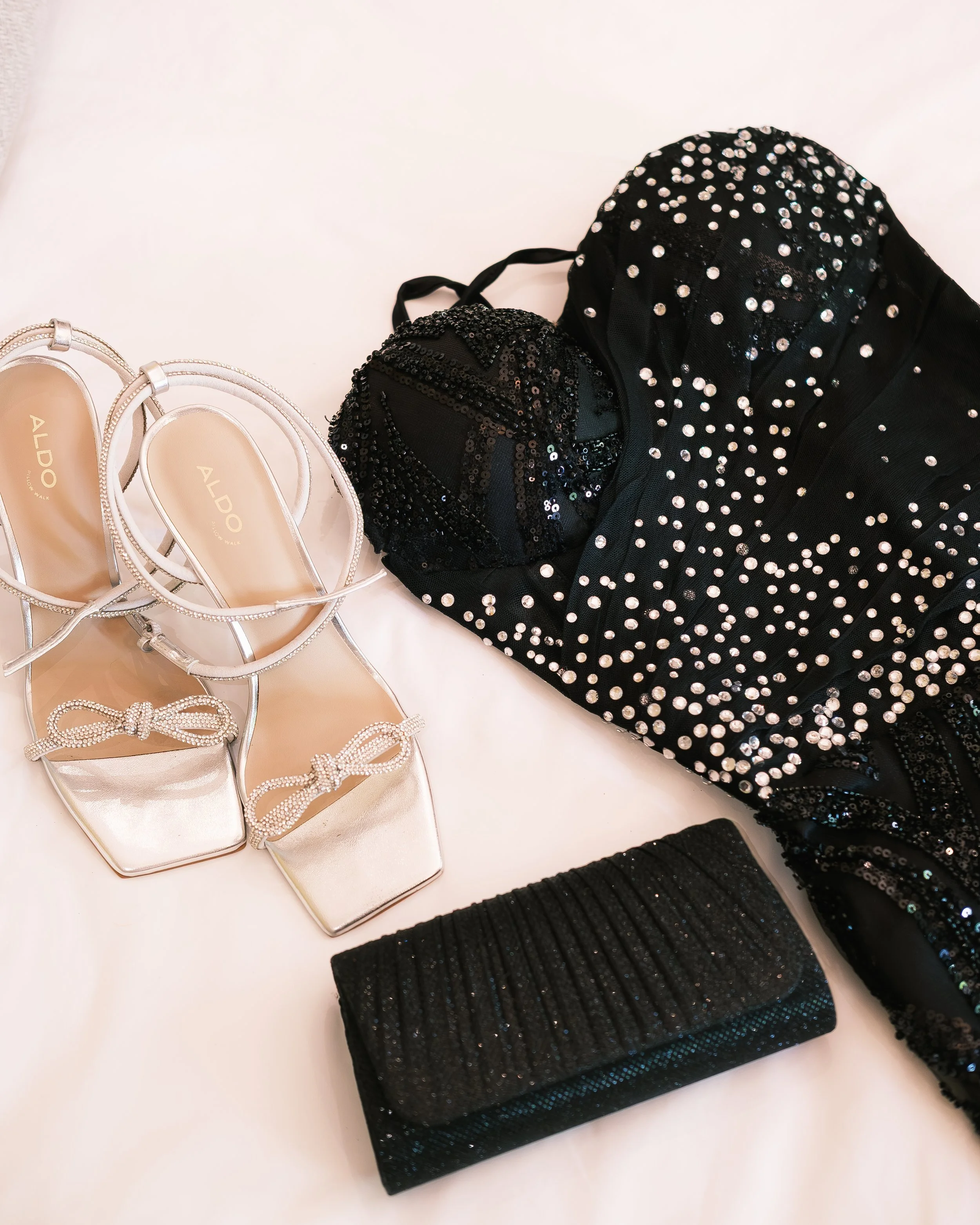 A pair of metallic silver high-heeled sandals with decorative bows, a black clutch with a sparkly finish, a black dress with silver embellishments, two black sequined headbands, and a black beaded clutch or wallet, all laid out on a white surface.